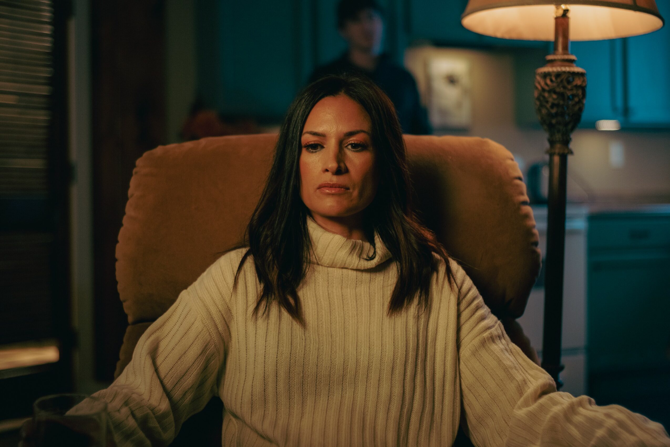 Woman with dark hair in a cream sweater sitting in a chair in a dimly lit room with a lamp behind her.