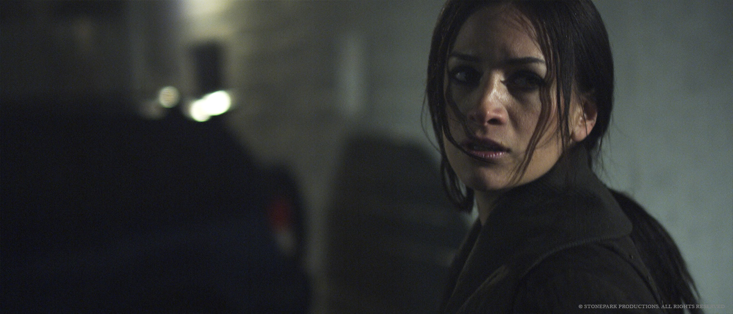 Woman with dark hair and dark clothing looks over her shoulder in a dimly lit setting.