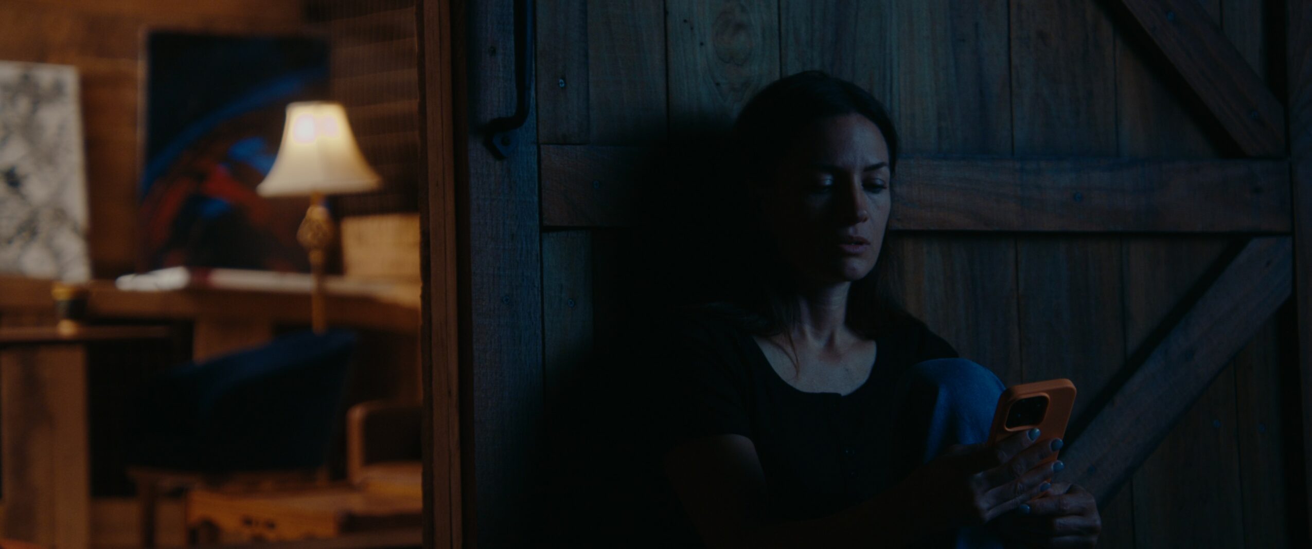 Woman sitting in a dimly lit room, looking at her phone, with a wooden wall behind her.