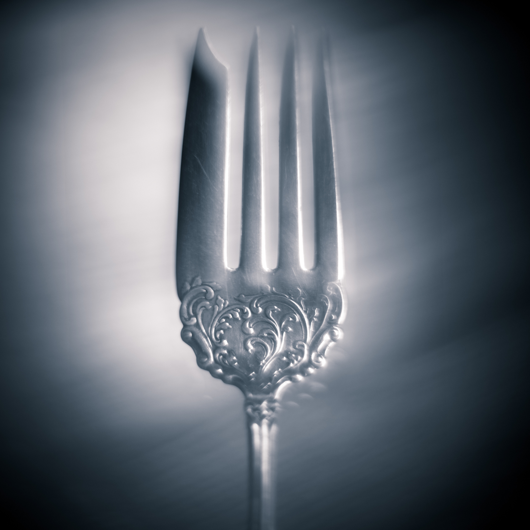 A silver fork with ornate handle, placed vertically on a reflective surface, with a blurred background.