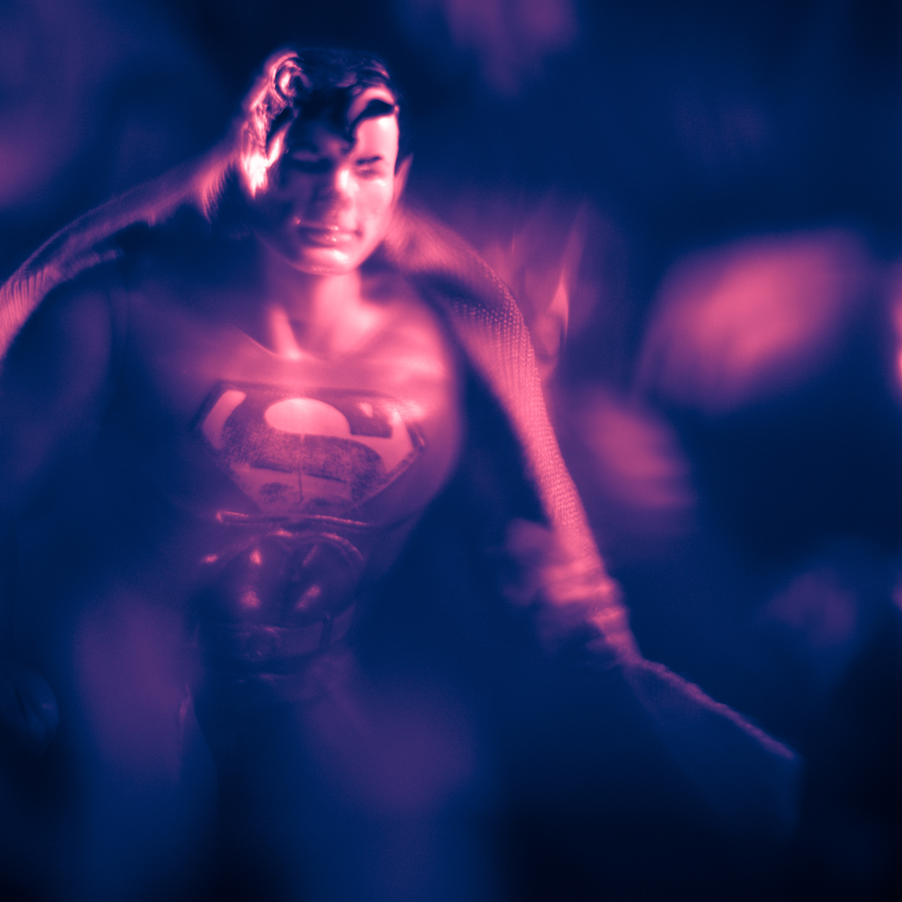 Supergirl in a dark setting with pink and purple lighting, wearing a Superman logo shirt, looking down with a hand on her head.
