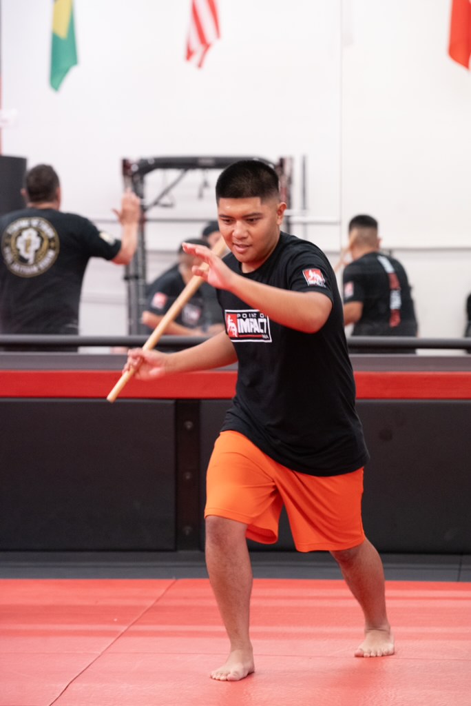 Young person practicing martial arts with a stick in a gym, wearing black shirt and orange shorts.