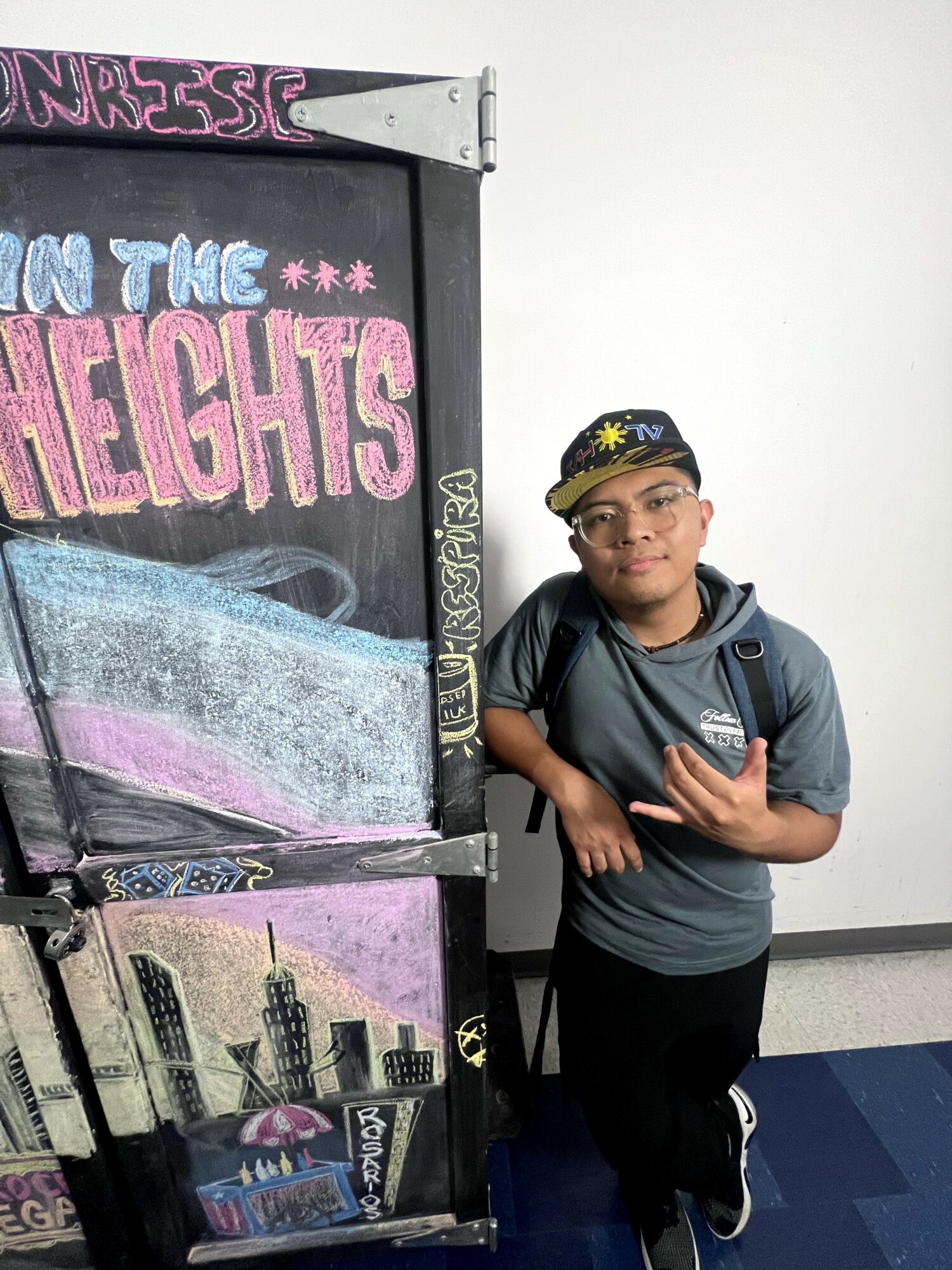 Young person standing next to a colorful sign with cityscape and text, holding a phone, wearing glasses, a cap, and a backpack.