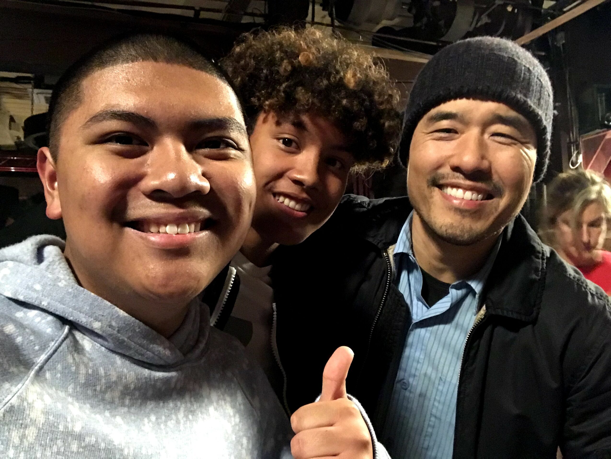 Three smiling people taking a selfie, two men and a woman, in a dimly lit indoor setting.