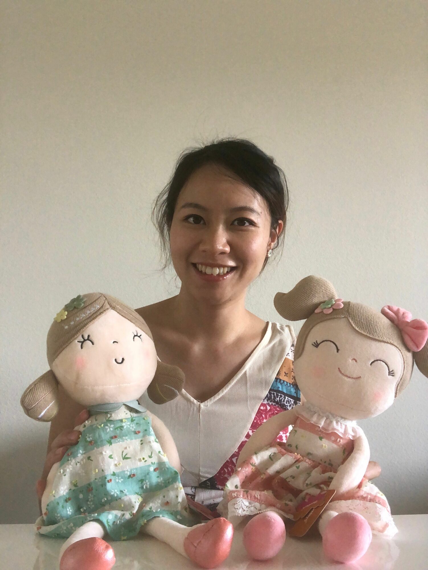 Smiling woman with two plush dolls, one wearing a striped dress, against a plain background.