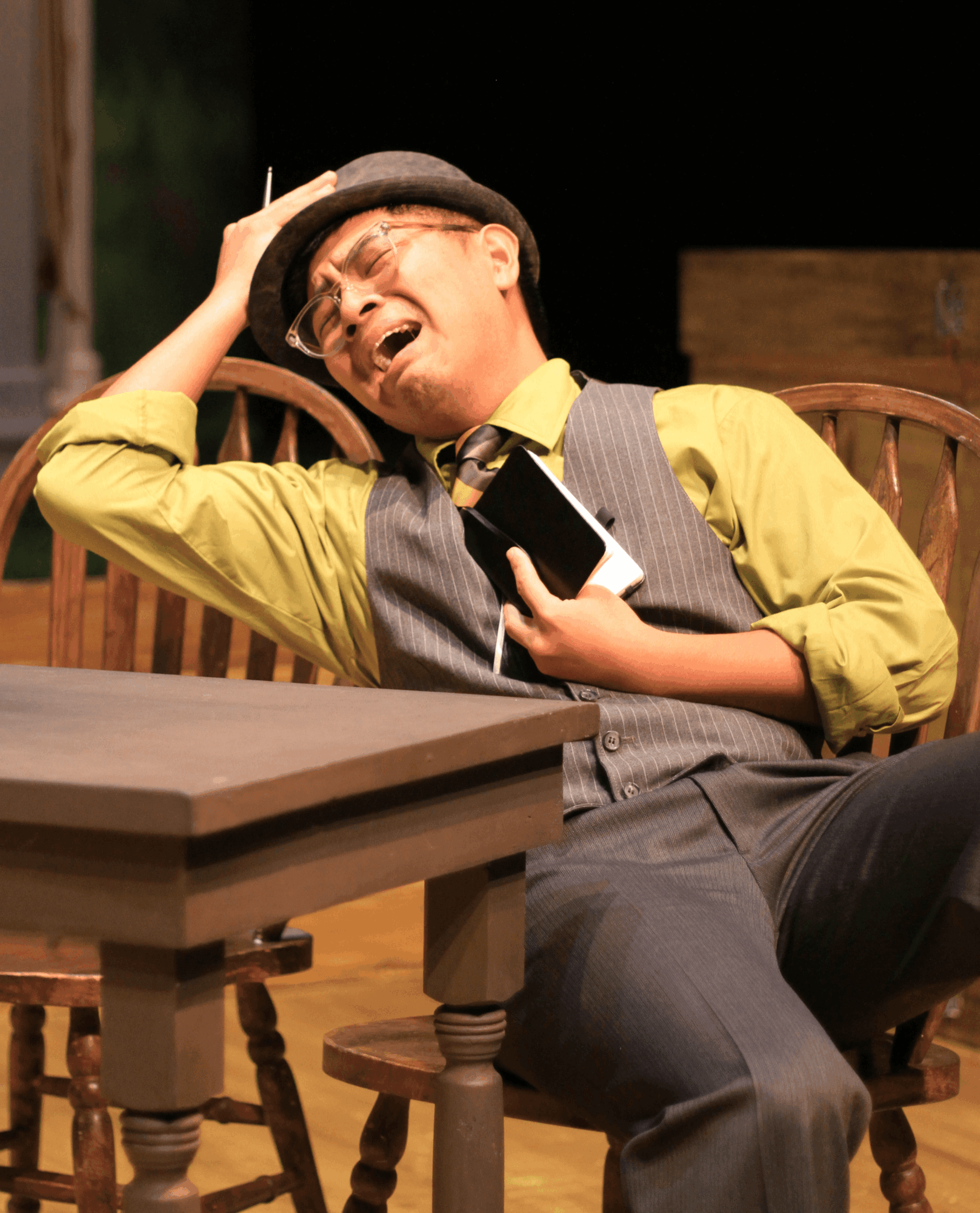 Person wearing a hat, yellow shirt, and gray vest, sitting on a wooden chair, laughing, holding a phone, in a dimly lit room.