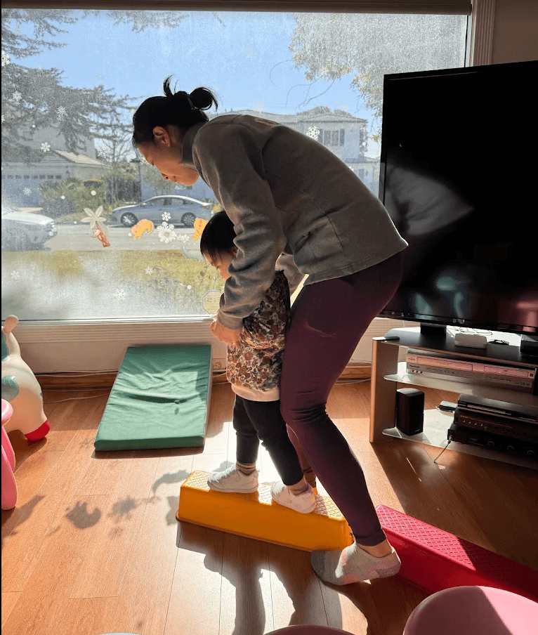 Adult and child practicing yoga on foam blocks indoors near a large window with outdoor view.