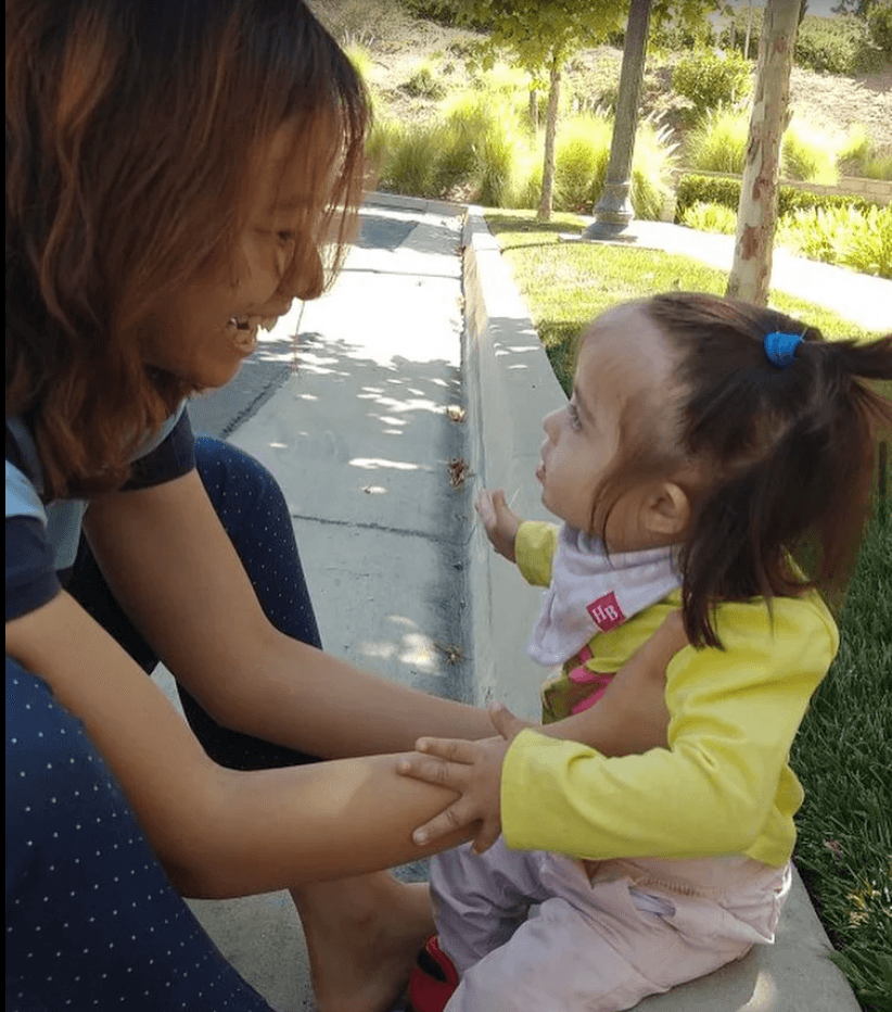 Adult and child holding hands and smiling outdoors on a sunny day, trees and a sidewalk in the background.