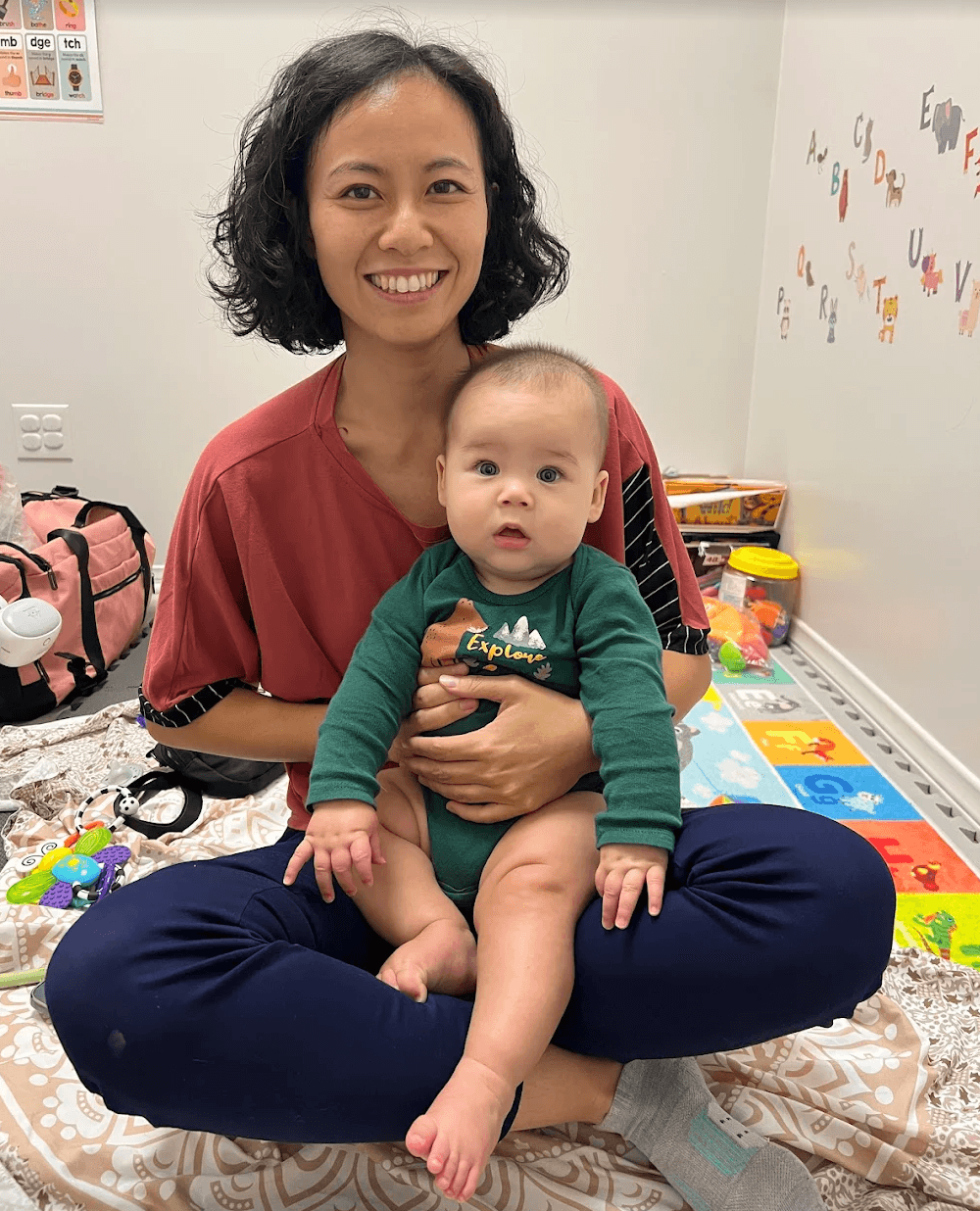 Woman sitting cross-legged holding a baby on her lap, smiling, in a colorful playroom with toys and wall decorations.