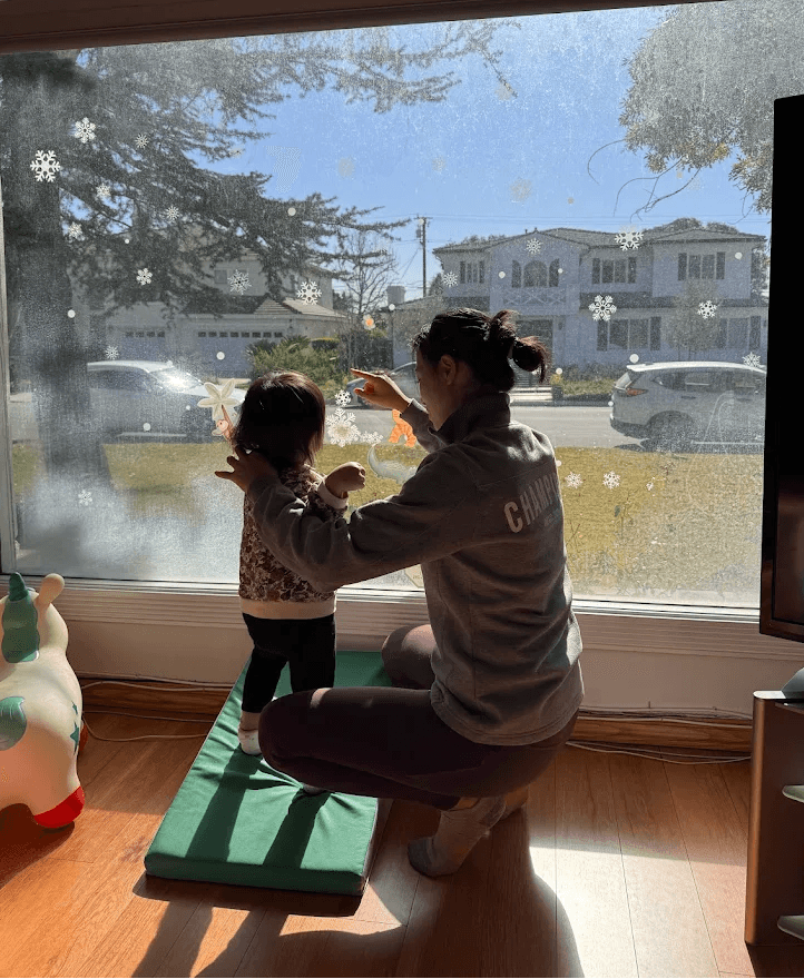 Adult and child looking out window with snowflake decorations, sunlight streaming in, and cars outside.