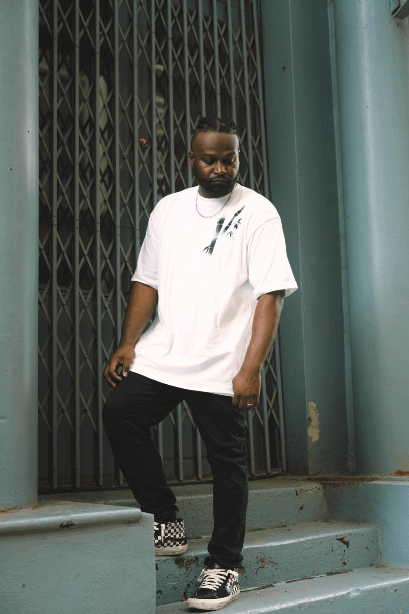 Young man with a beard stands on steps in front of a metal gate, wearing a white oversized t-shirt, black pants, and checkered shoes.