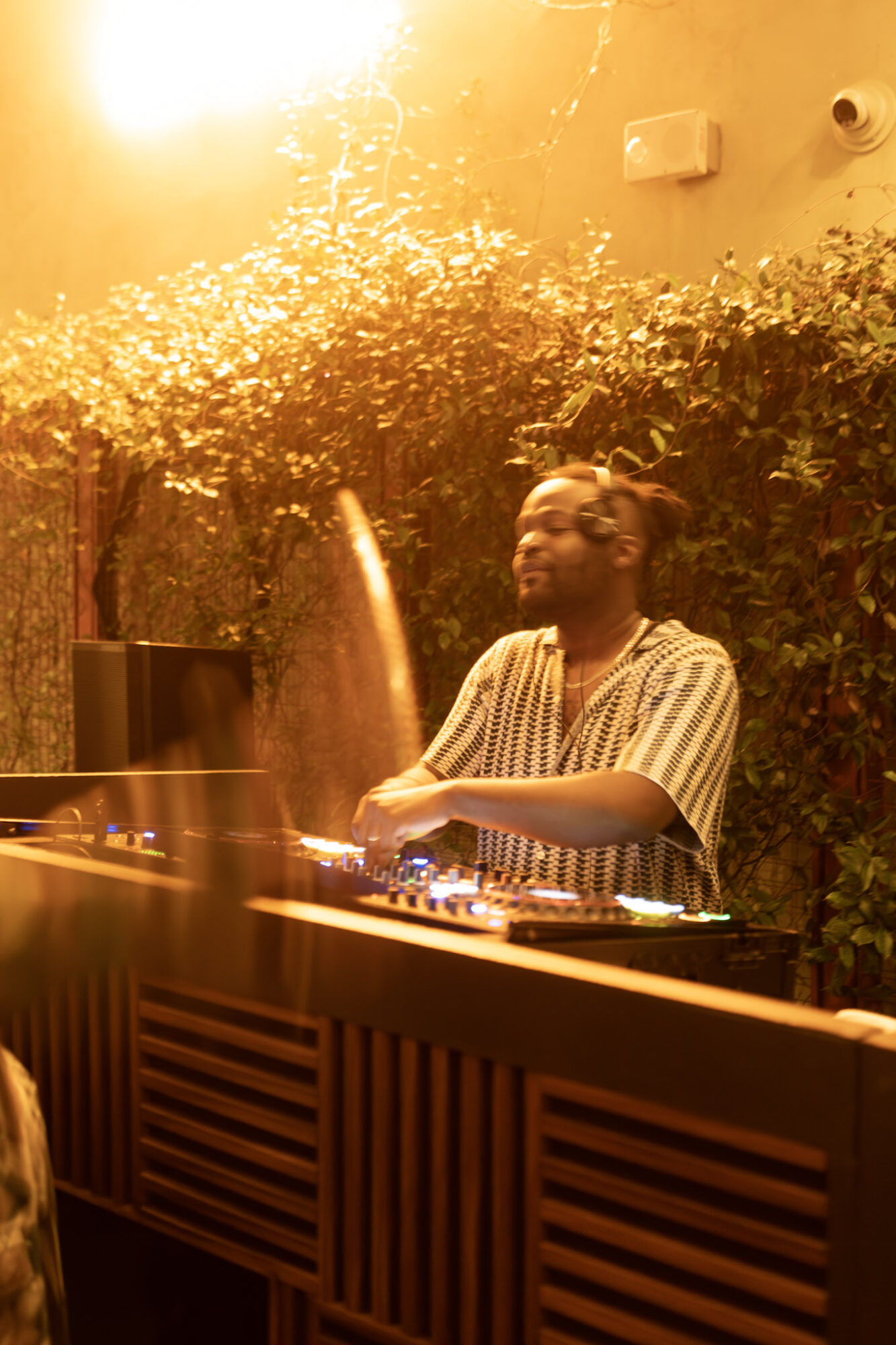 Person DJing outdoors with sunlight and greenery, wearing striped shirt, smiling, with DJ equipment in front.