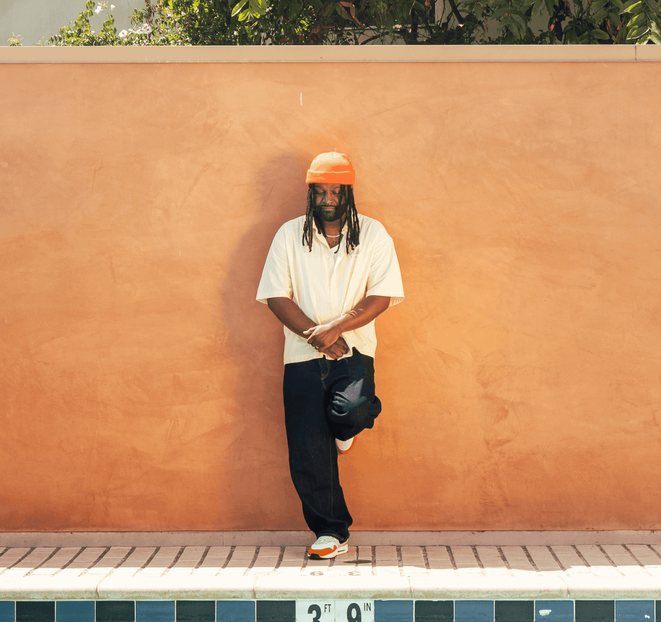 Person standing against a peach-colored wall, wearing an orange hat, white shirt, and dark pants, with one foot resting on the wall.