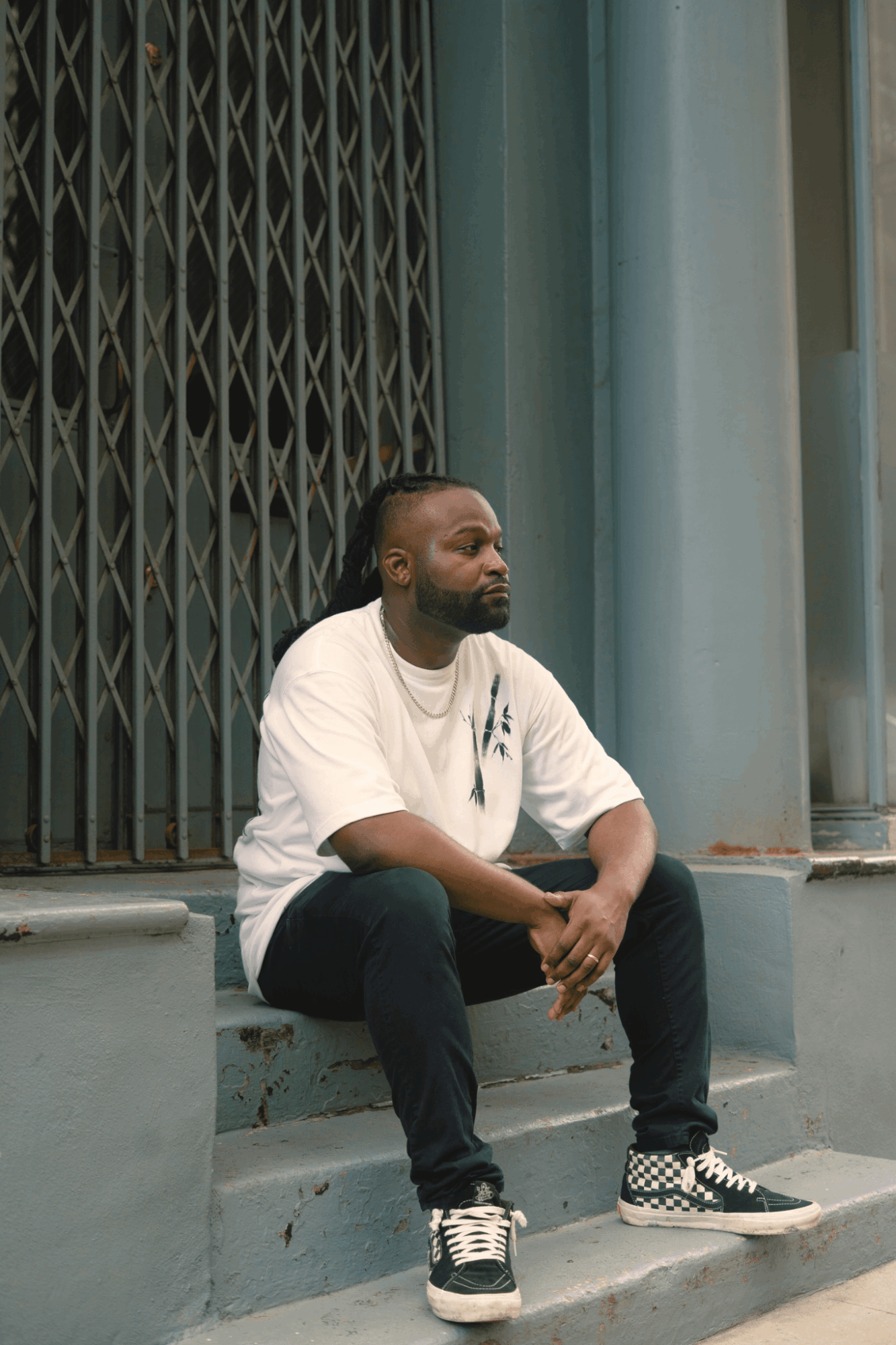 Man sitting on steps in front of a metal gate and blue wall, wearing a white t-shirt, black pants, and checkered shoes.