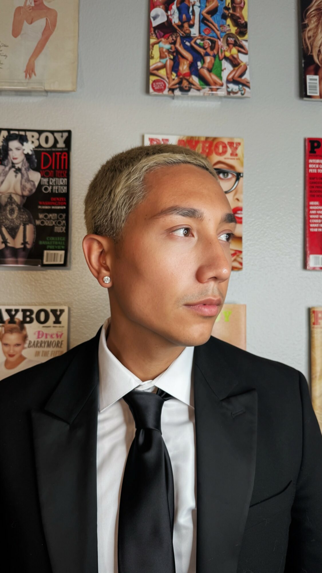 Young person in a black suit and tie looking to the right, with magazine covers on the wall behind them.