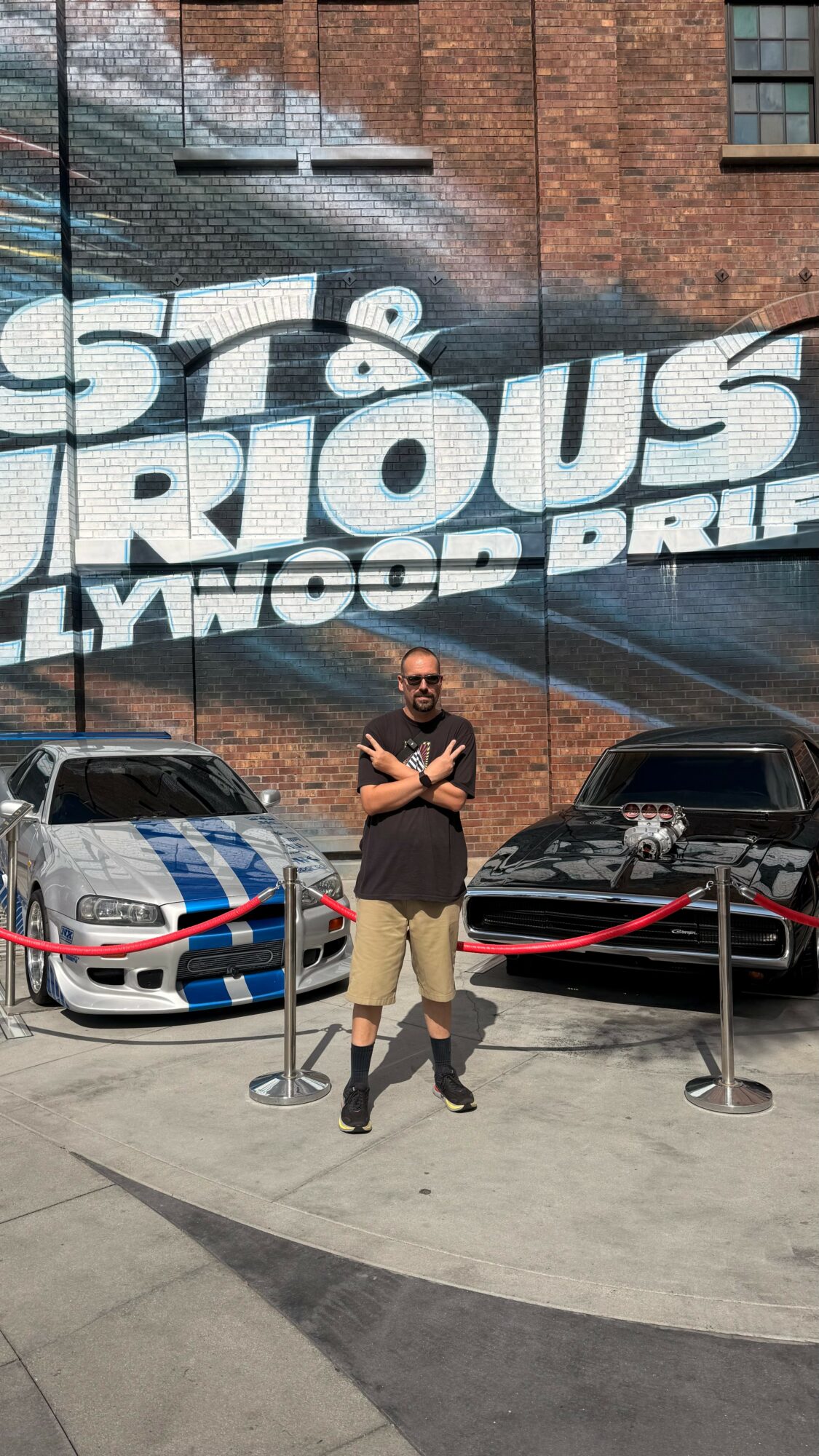Person standing in front of a mural with cars parked behind, arms crossed, wearing sunglasses and a black shirt.