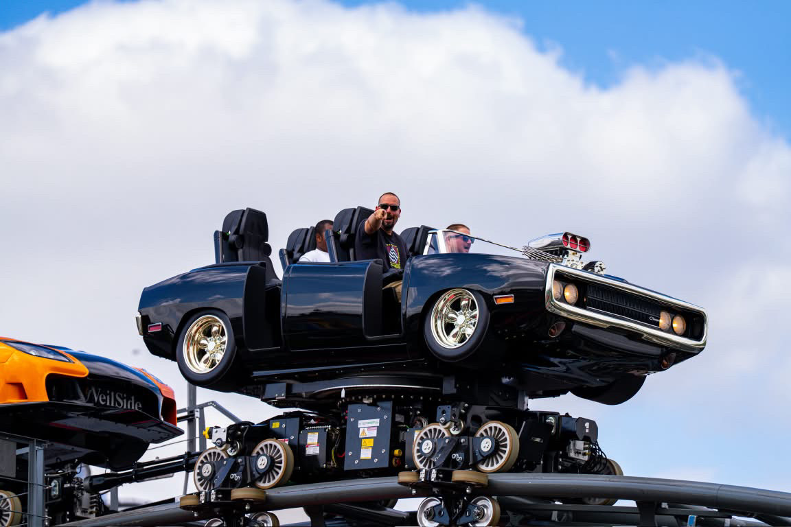 Person sitting in a black convertible car on a moving platform, with a blue sky and clouds in the background.