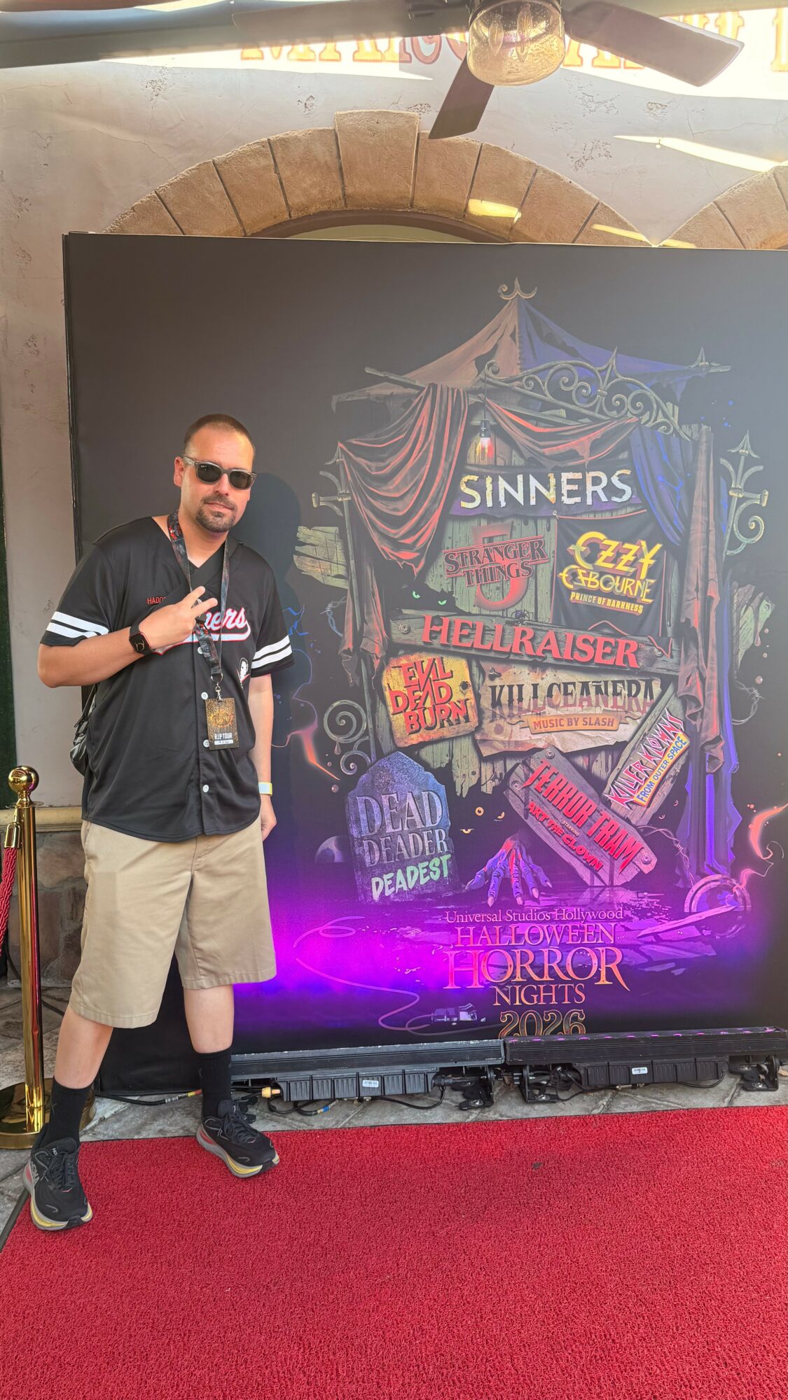 Man standing next to a colorful chalkboard sign with event details, on a red carpet, outdoors, with a brick arch above.