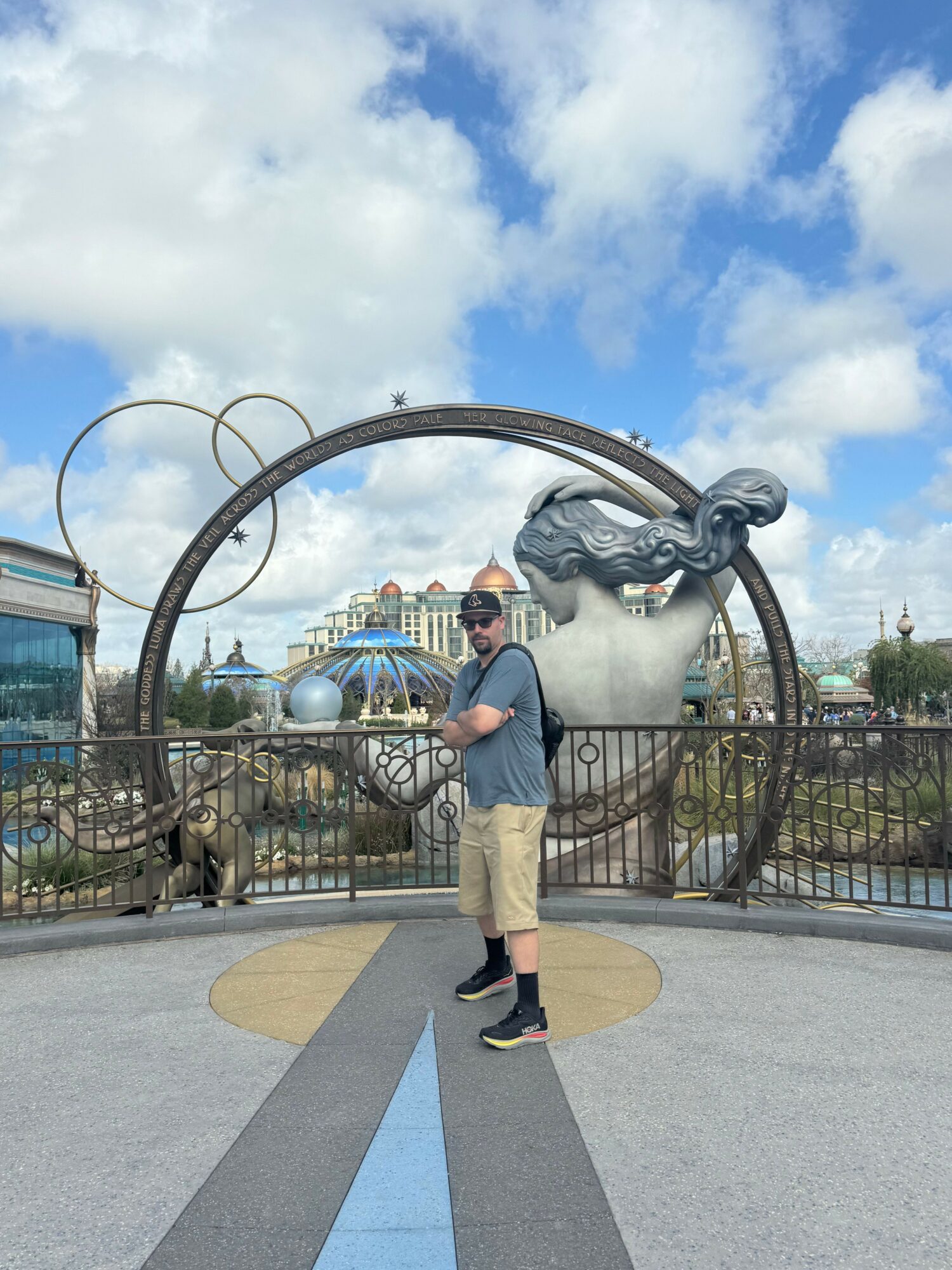 Person walking past a large, abstract metal sculpture outdoors with a cloudy sky background.
