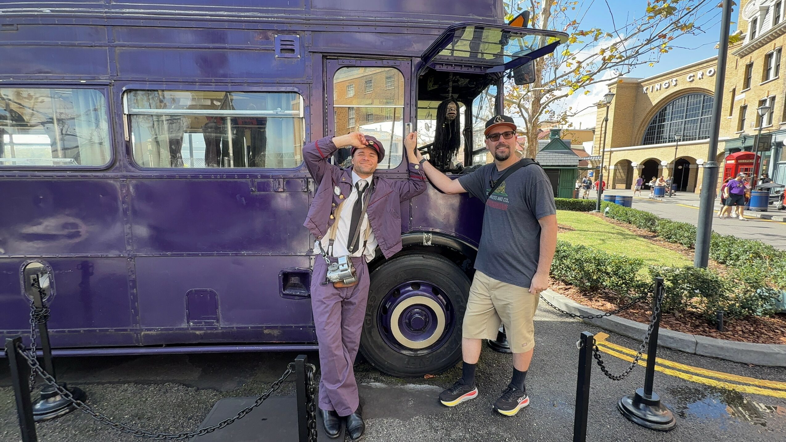 Two people stand in front of a purple bus, one waving and the other with hand on the bus, in an outdoor urban setting.