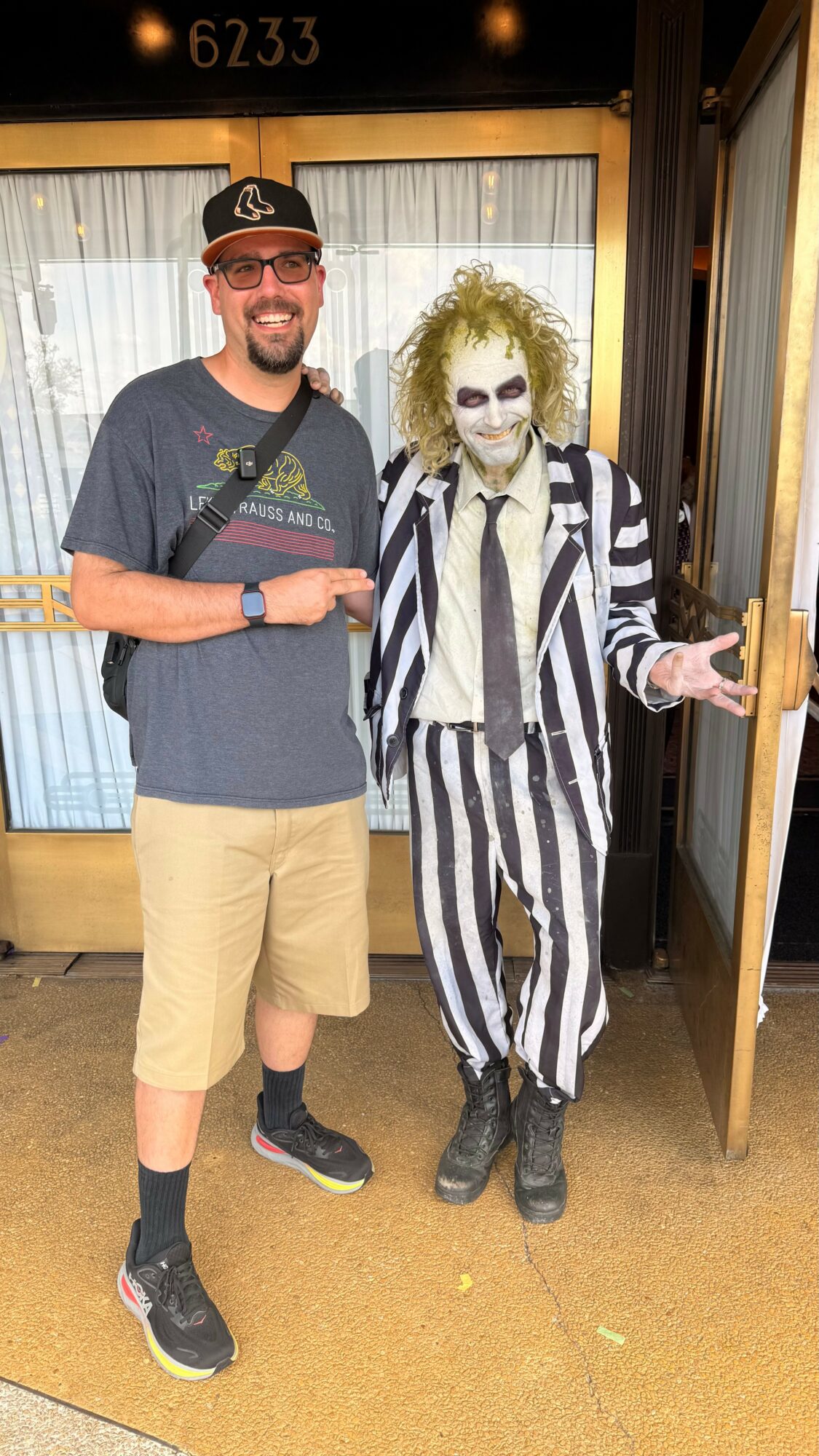 Person with a beard and glasses smiling, wearing a baseball cap, t-shirt, shorts, and sneakers, standing next to a person dressed as Beetlejuice in striped suit and makeup, inside a building.