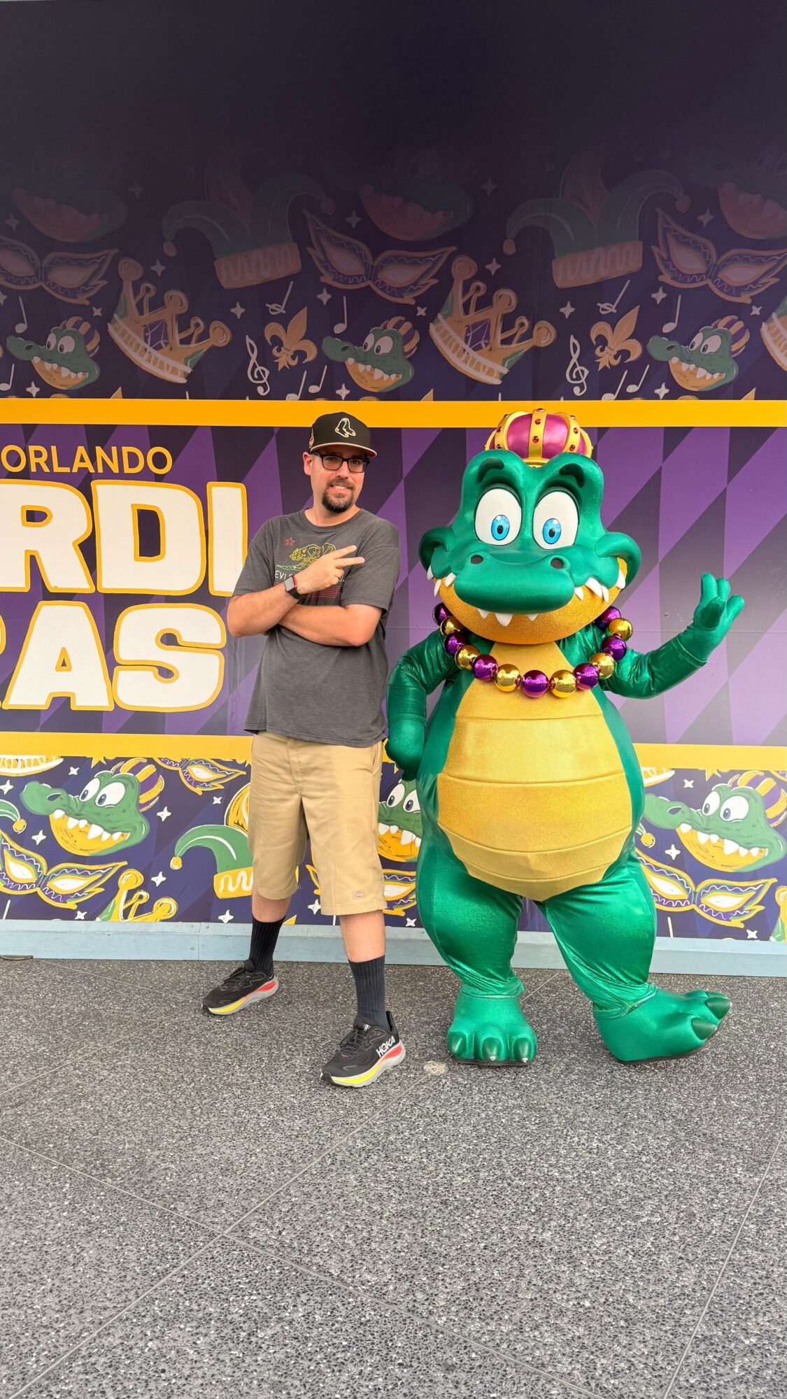 Person standing next to a large green dinosaur mascot with a crown and jewelry, in front of a colorful backdrop.