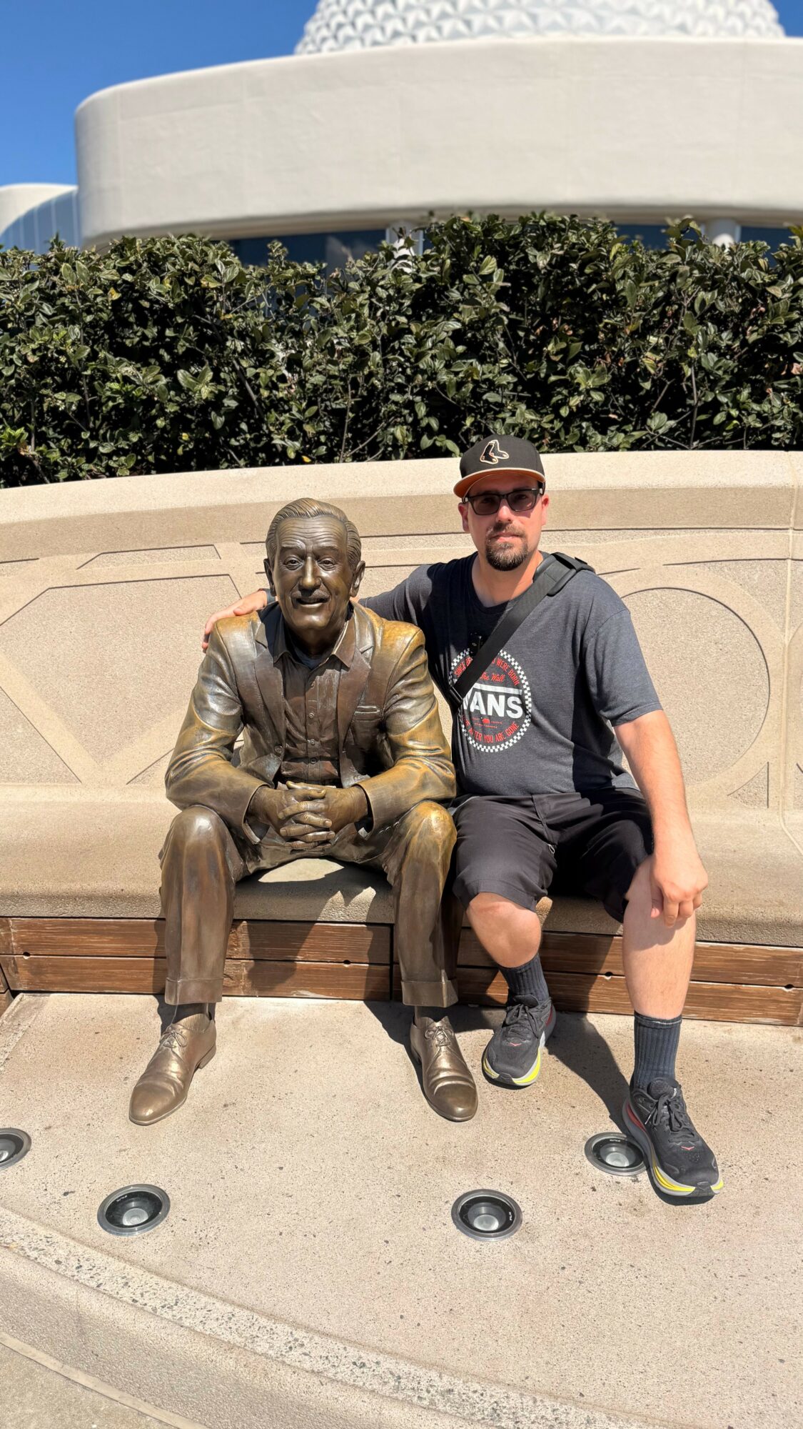 Person sitting next to a bronze statue of a man in a suit on a park bench, with greenery and a building in the background.