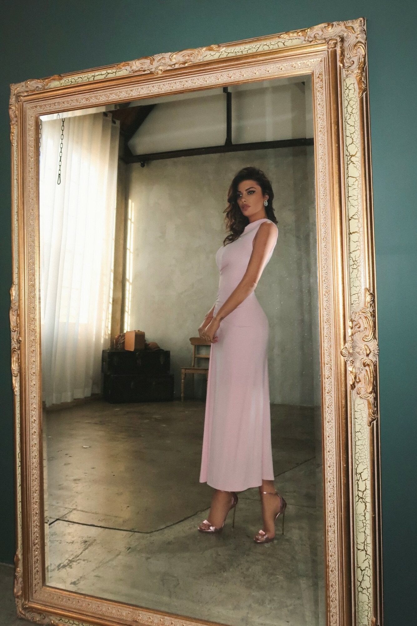 Woman in a long pink dress standing in front of a large ornate mirror, reflection shows her full body, room background.