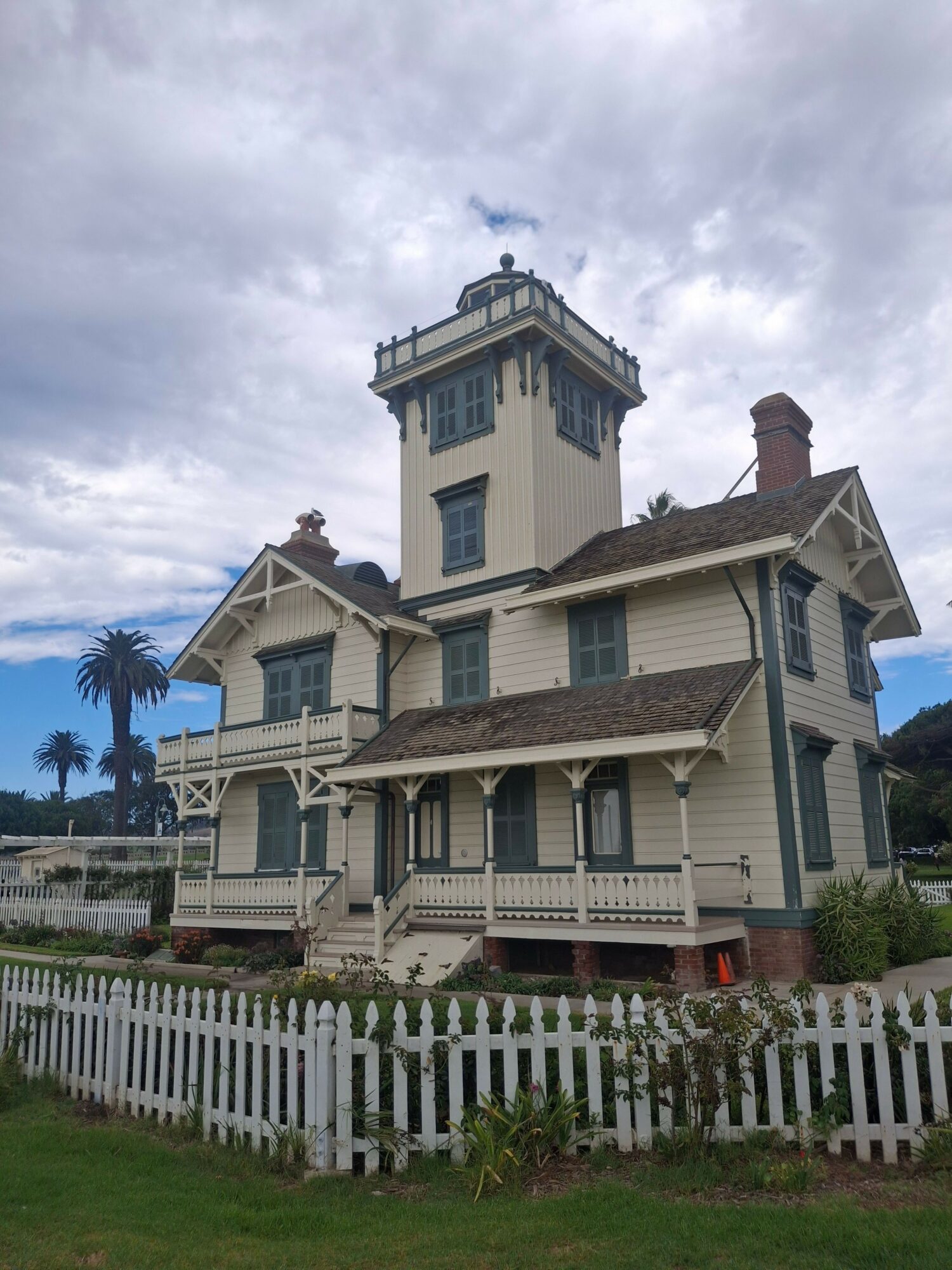 Victorian-style house with a white picket fence and a garden, under a cloudy sky.