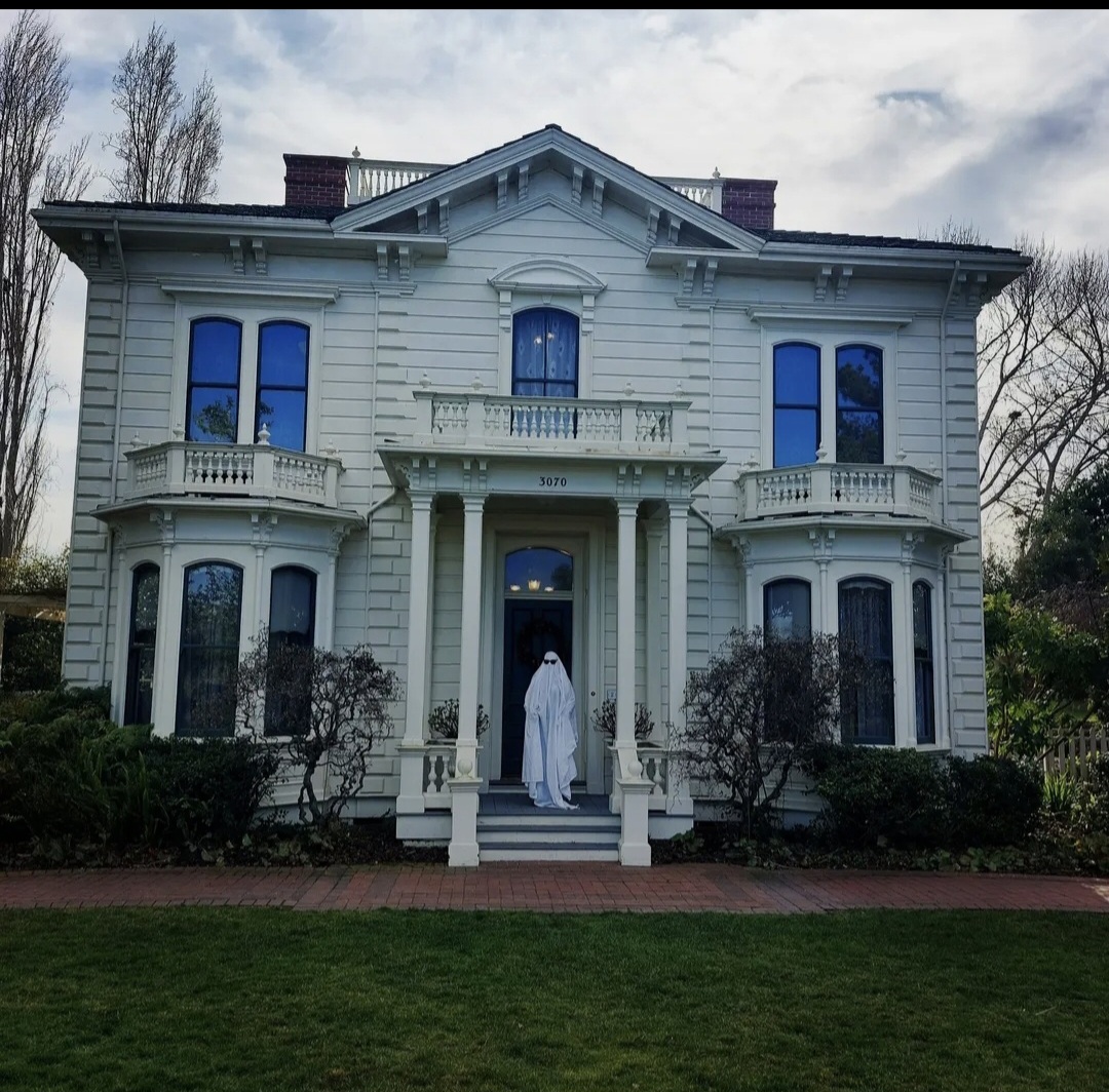 Large white Victorian-style house with a person standing on the front steps, trees in the background, cloudy sky.