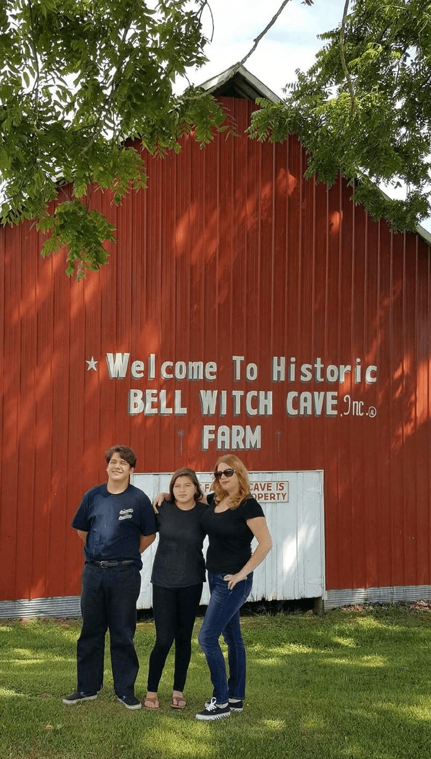 Three women stand in front of a red barn with a sign welcoming visitors to Bell Witch Cave and farm.