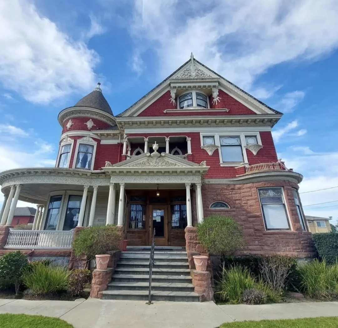 Victorian-style red house with a turret, front porch, and stairs, under a partly cloudy sky.
