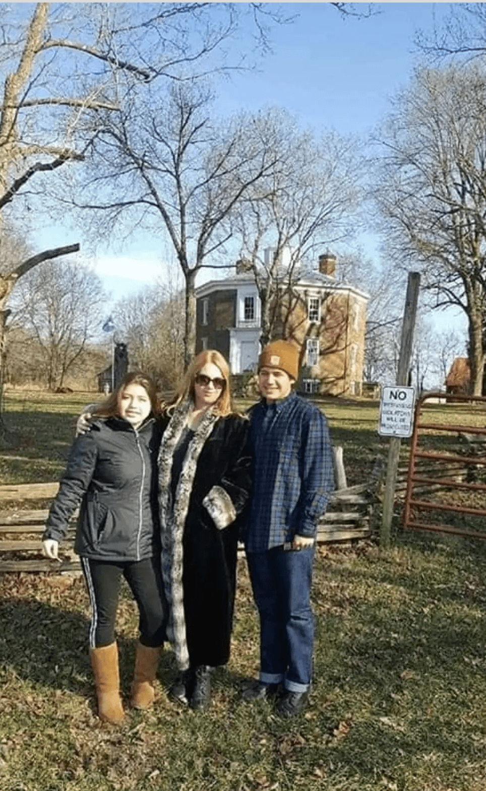 Three people standing outdoors in front of a house with trees, one wearing sunglasses and a hat, sunny day.