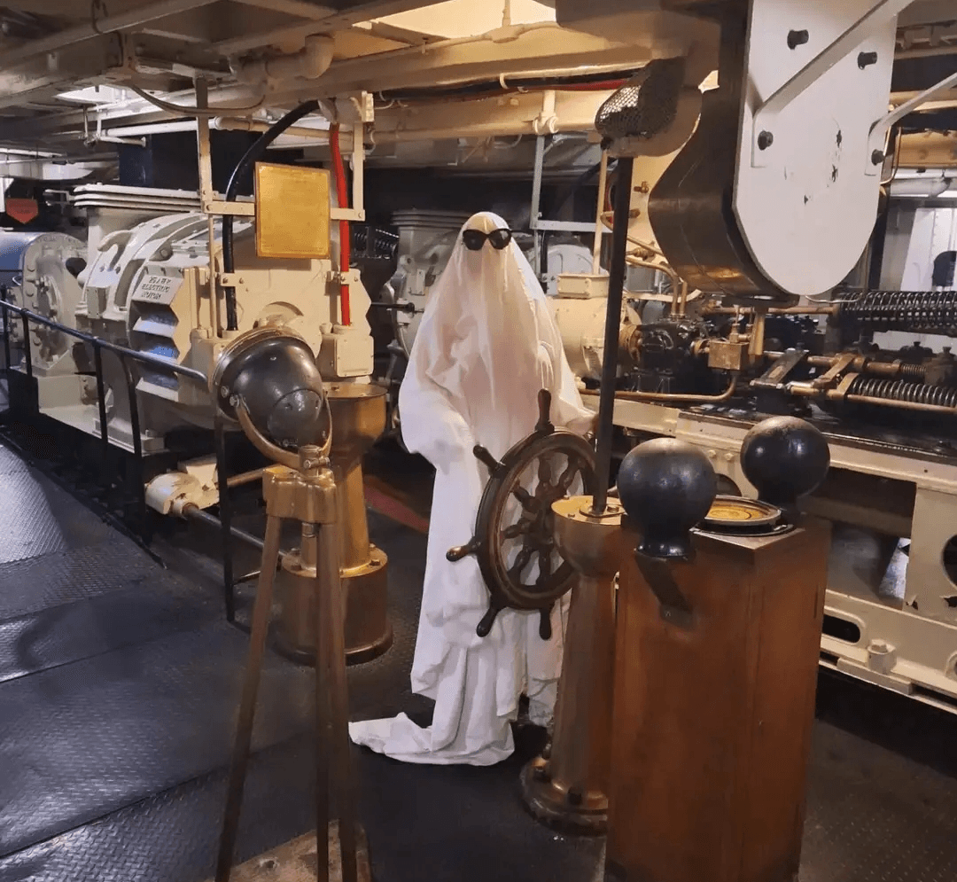 Person dressed as a ghost with sunglasses standing among ship machinery and nautical equipment.
