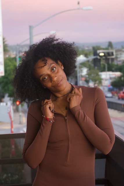 Young woman with curly hair leaning on a railing outdoors during sunset, wearing a brown long-sleeve top, looking at the camera.