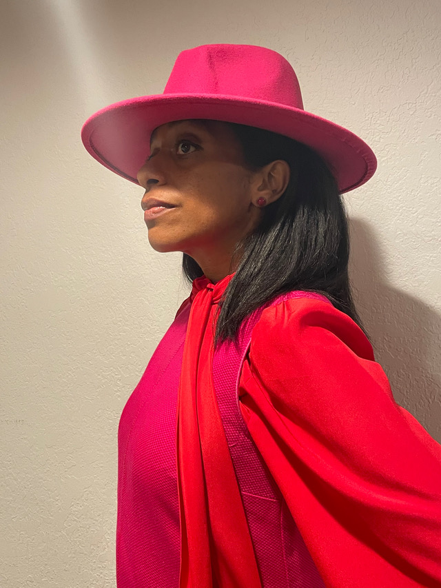 Woman wearing a large pink hat and red outfit, looking to the side, against a plain background.