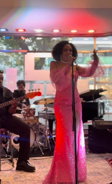 A woman in a pink dress sings into a microphone on stage with musicians playing instruments behind her.