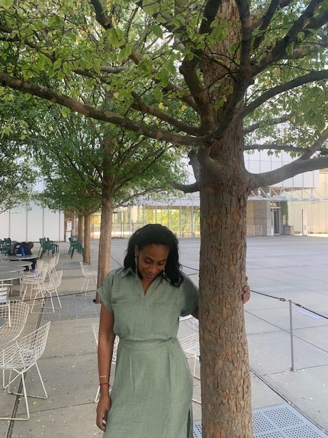 Woman standing next to a tree on a sidewalk, with buildings and a bench in the background.