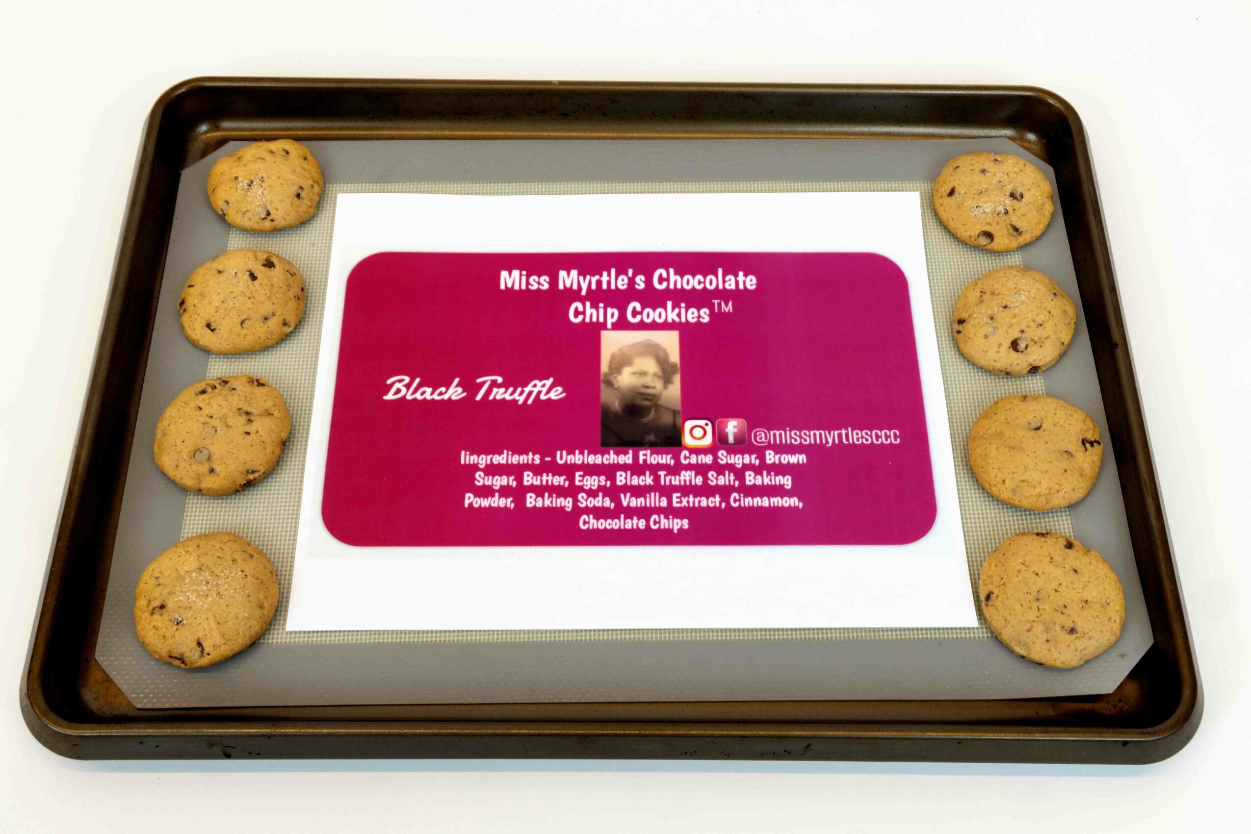 Baking tray with eight chocolate chip cookies and a pink label in the center, on a white surface.