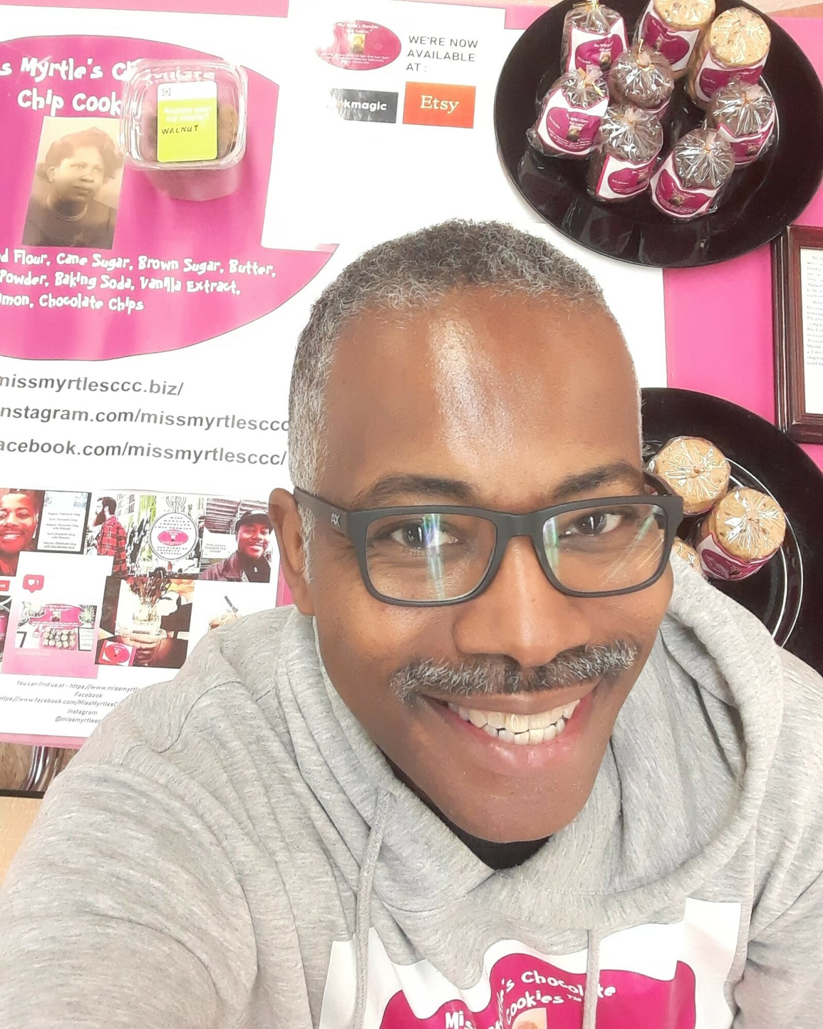 Man with glasses smiling in front of pink display with cupcakes and promotional materials.