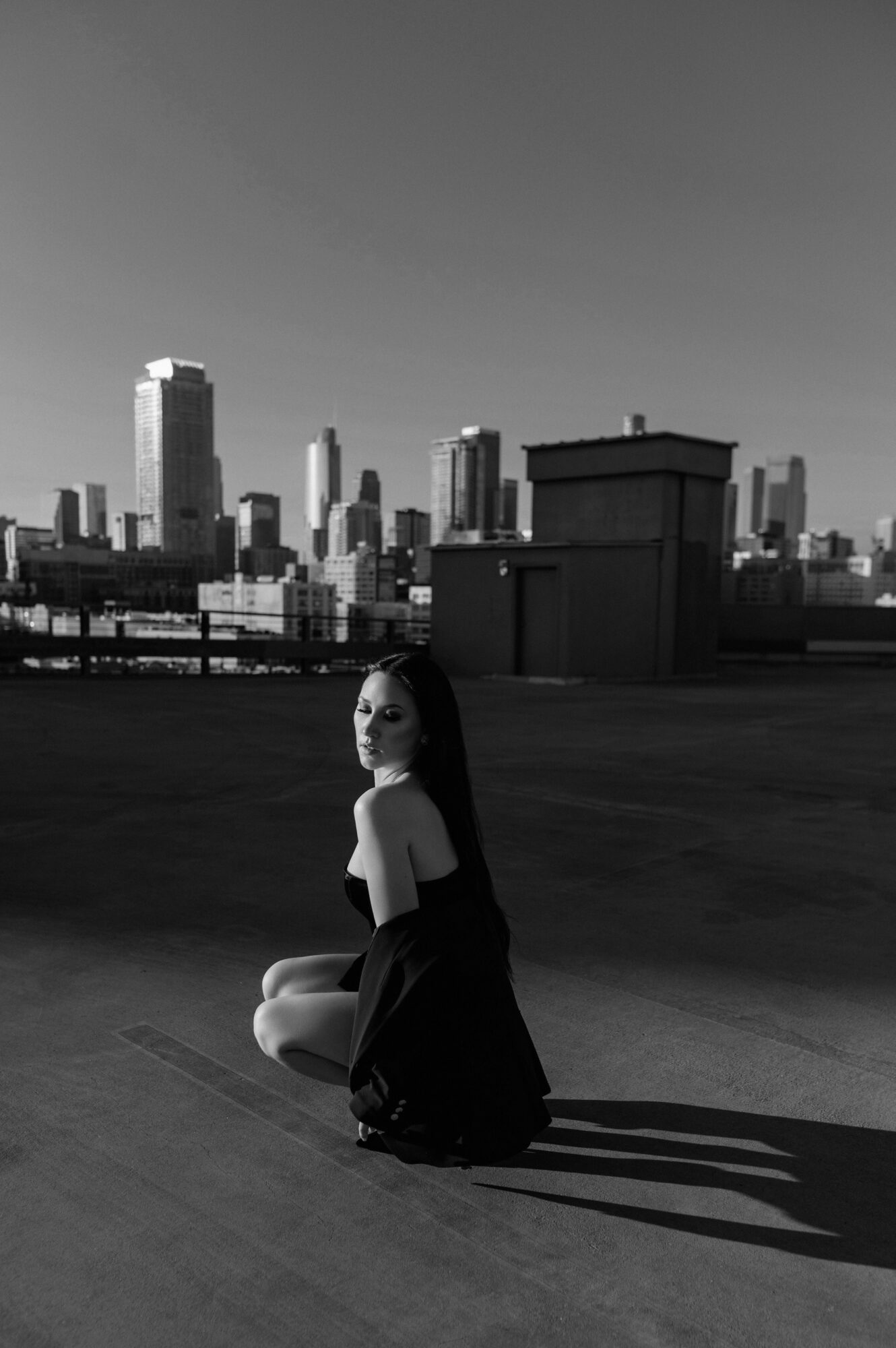 Woman crouching on rooftop with city skyline in background, shadows cast on ground, black and white image.