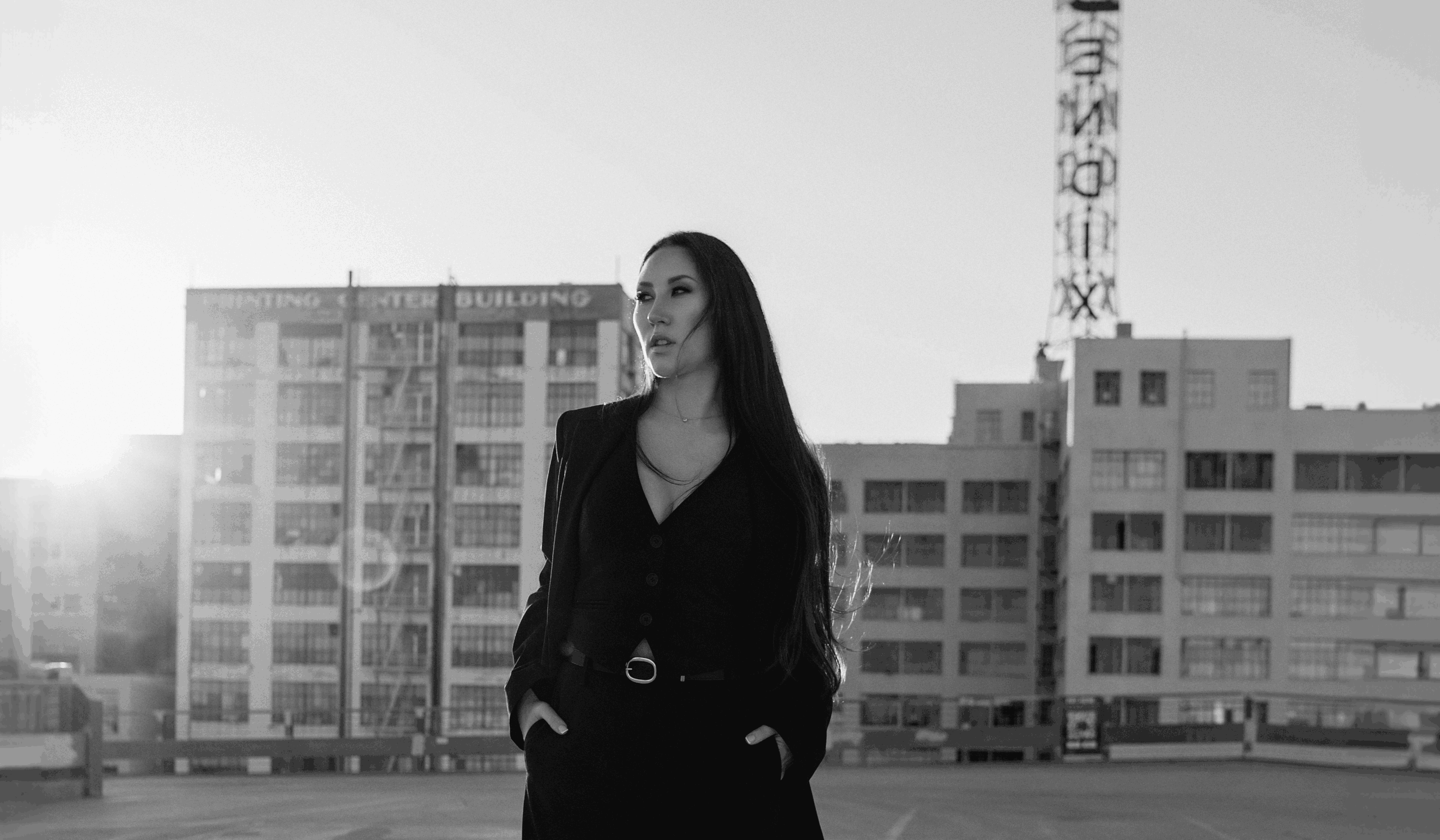 Woman with long dark hair standing outdoors in front of buildings, sunlight behind her, wearing a dark outfit with hands in pockets.