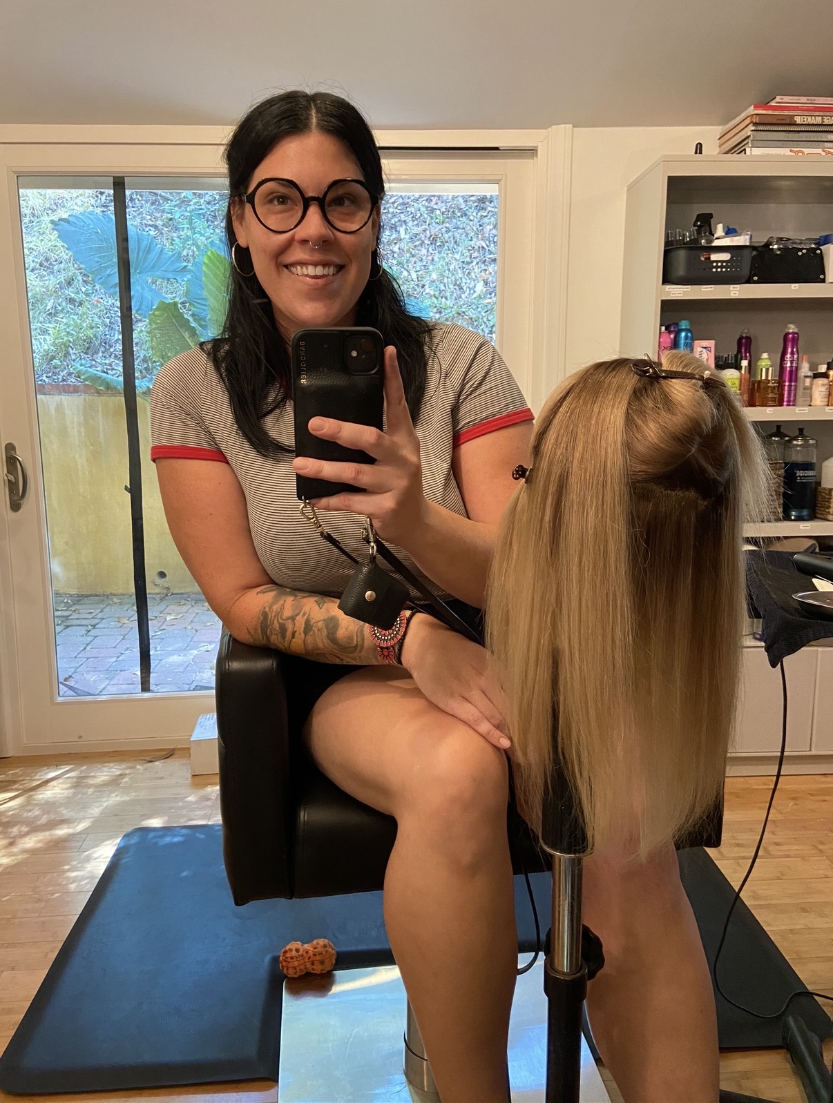 Woman with glasses smiling, holding a mannequin head with long blonde hair, in a room with a sliding glass door and shelves.