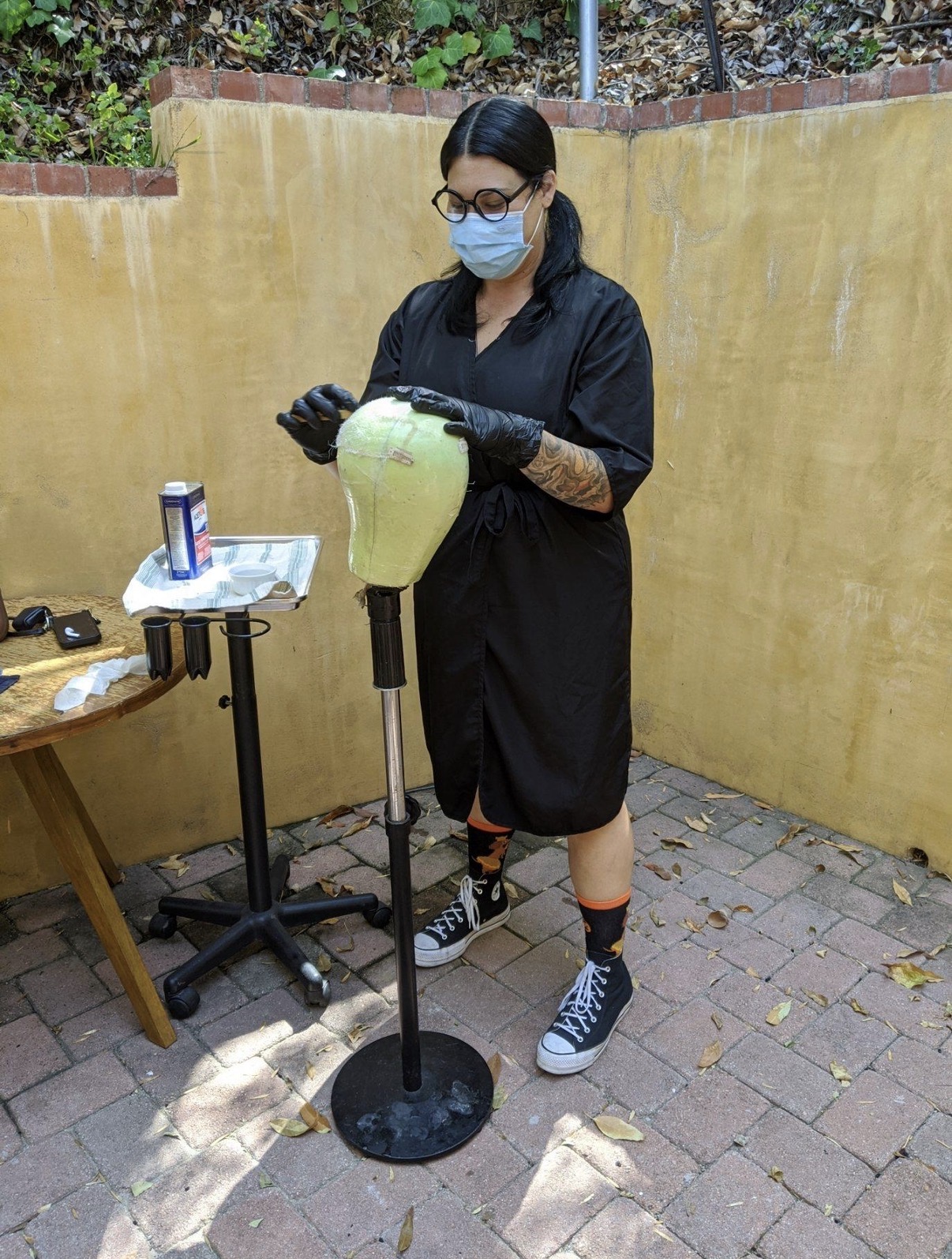 Person wearing a face mask and gloves, working with a green object on a stand outdoors, with a yellow wall behind.