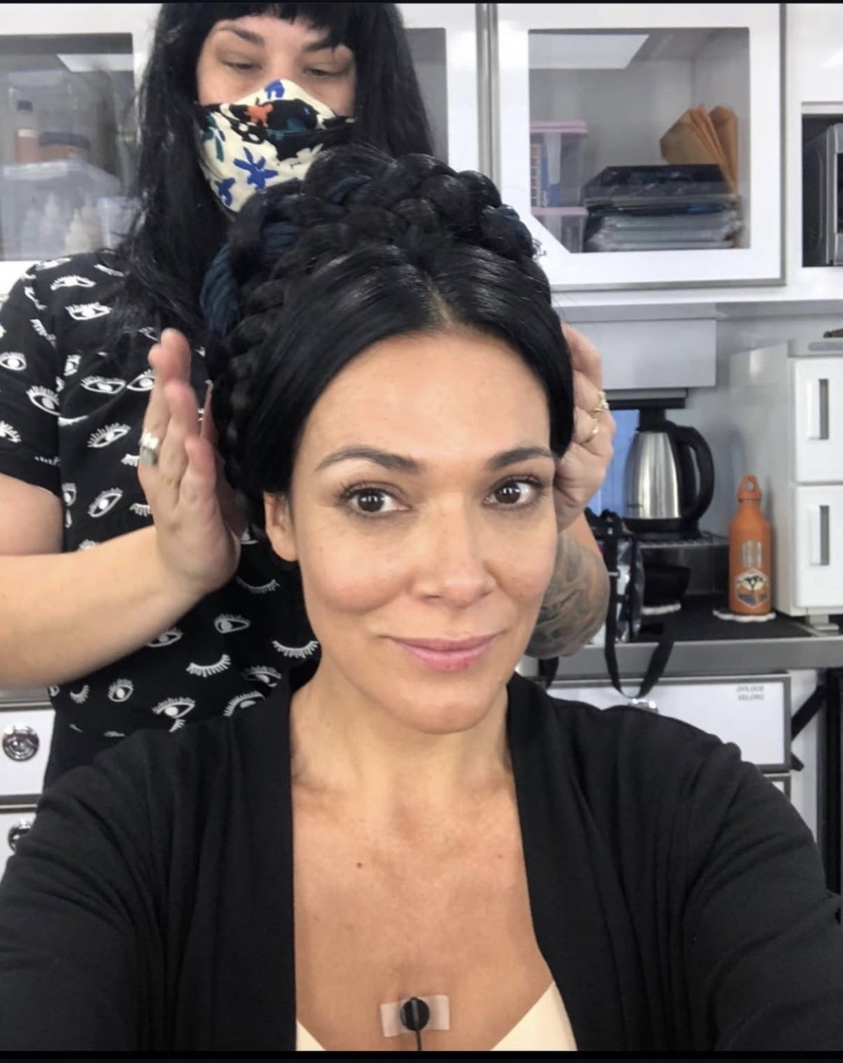 Woman with styled hair taking a selfie in a kitchen, person with a mask working on her hair behind her.