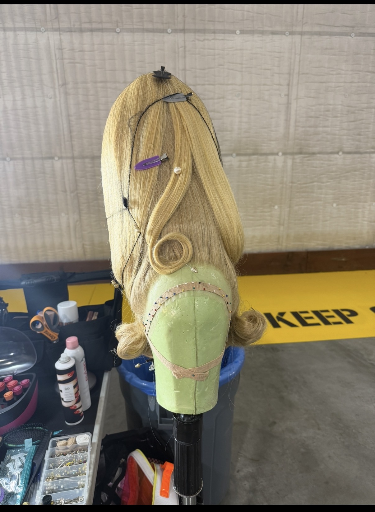 Blonde mannequin head with curled hair, hair clips, and a green foam block for styling practice, in a workshop setting.