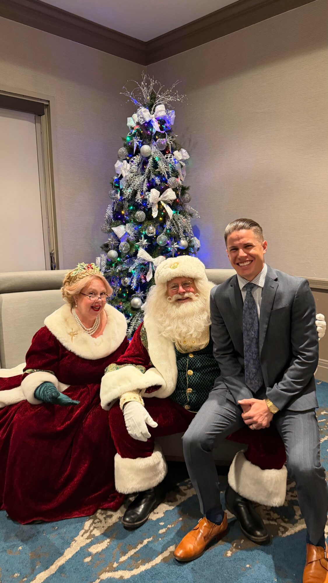A man in a suit sitting next to Santa Claus and a woman dressed as Mrs. Claus, with a decorated Christmas tree in the background.