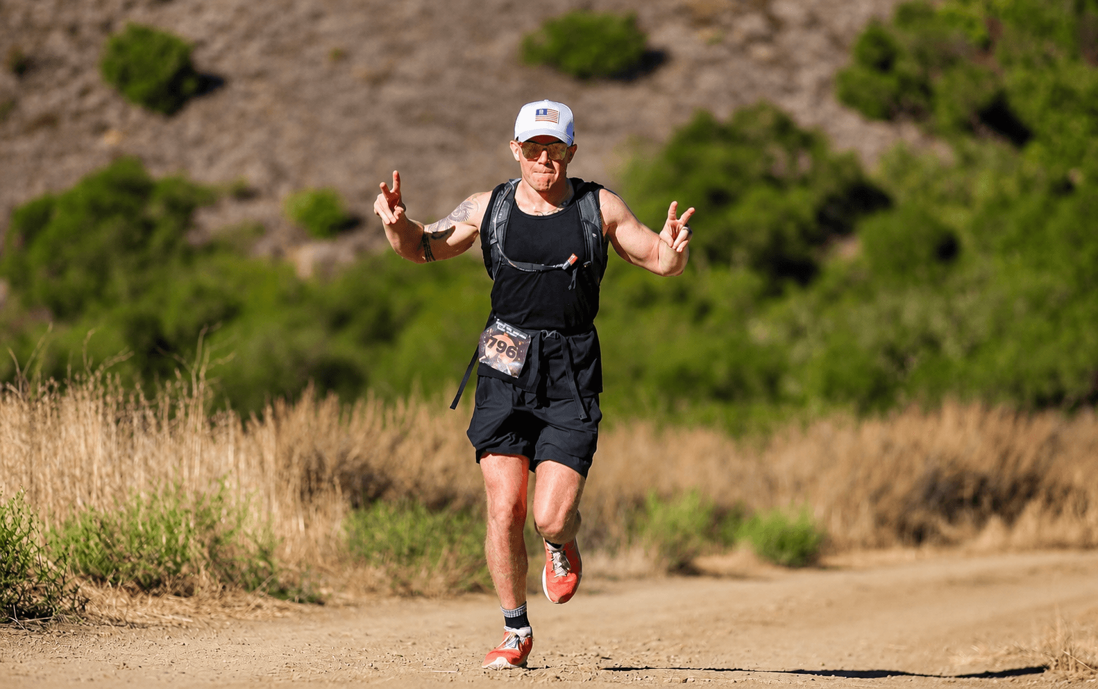 Person running on a dirt trail in a natural landscape with grass and trees, wearing a cap, sunglasses, and athletic clothing.
