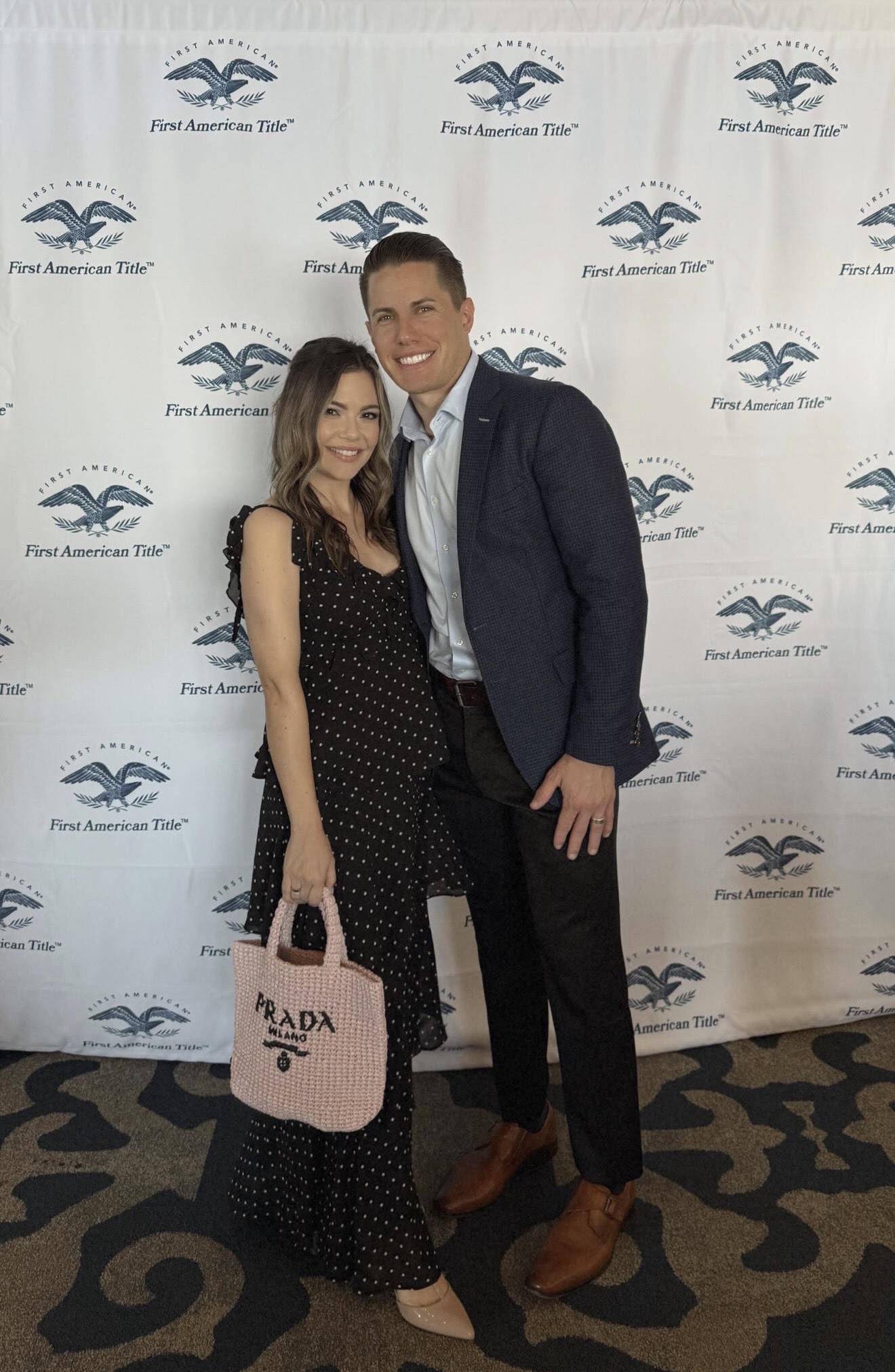 Two people standing in front of a backdrop with a logo, smiling, one holding a pink bag, both dressed formally.