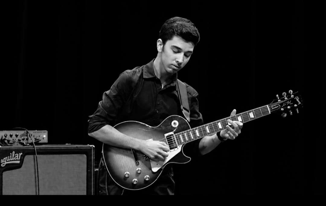 Young man playing an electric guitar on stage with a black background, focused on his instrument.